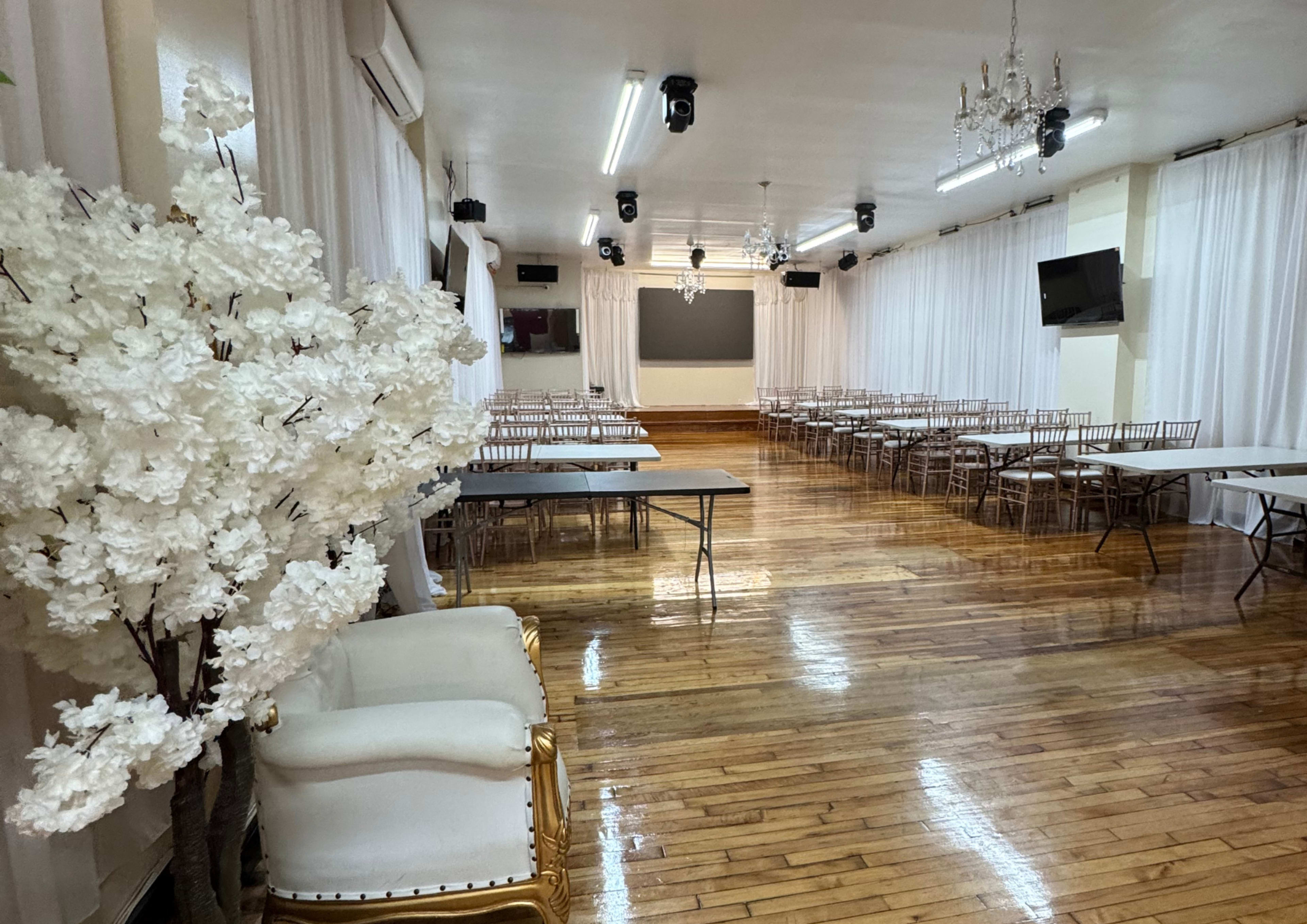 The image shows a large, empty indoor event space featuring rows of tables and gold-accented chairs, with white drapes and a decorative artificial tree.