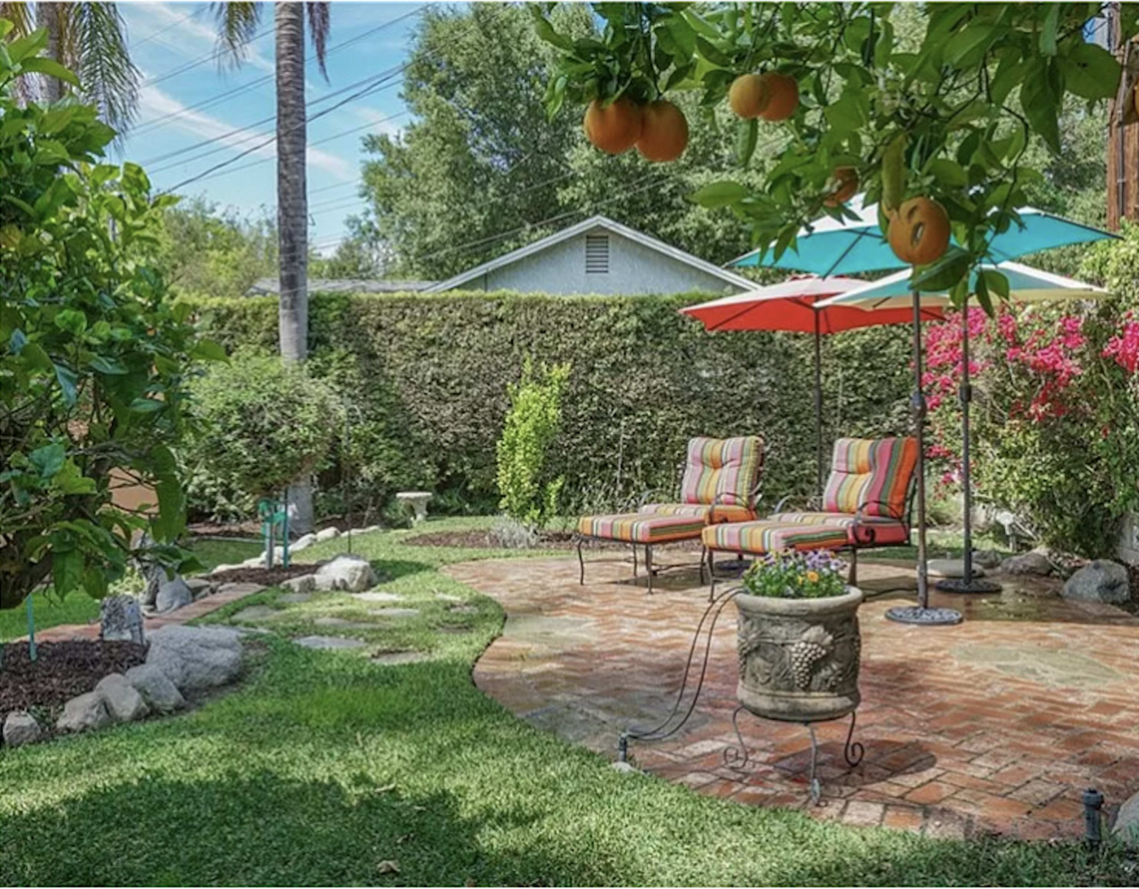 The image shows a backyard with two striped lounge chairs under an umbrella, surrounded by lush greenery and flowering plants.