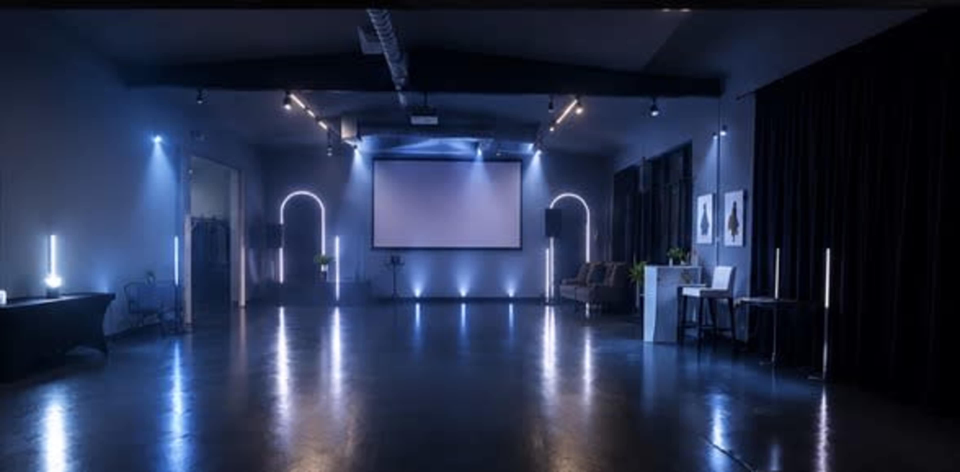 The image shows a dimly lit event space featuring a large screen, seating arrangements, and illuminated vertical light fixtures.