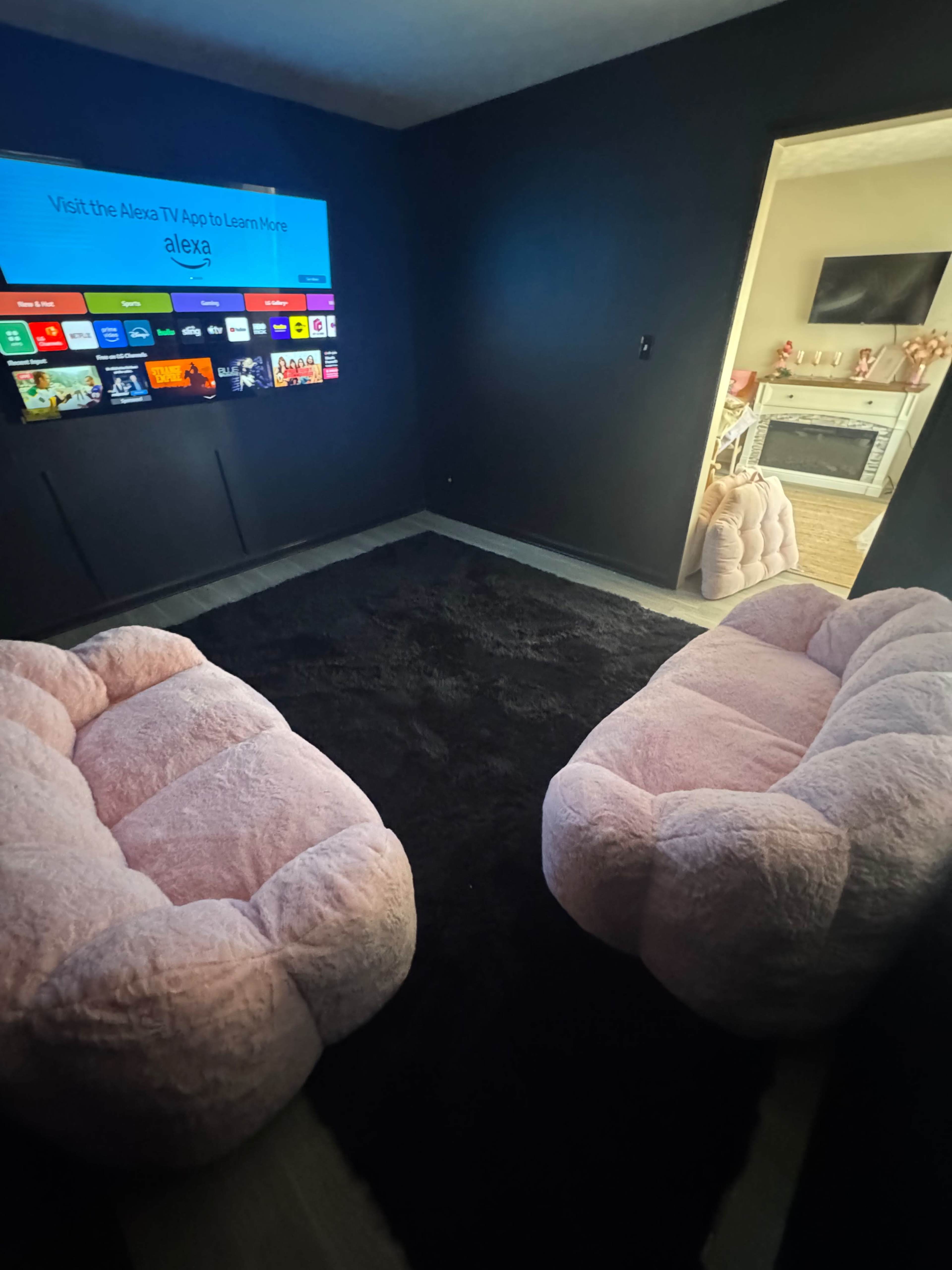The image shows a cozy room featuring two pink, fluffy couches facing a large television mounted on a dark wall, with a black rug covering the floor.