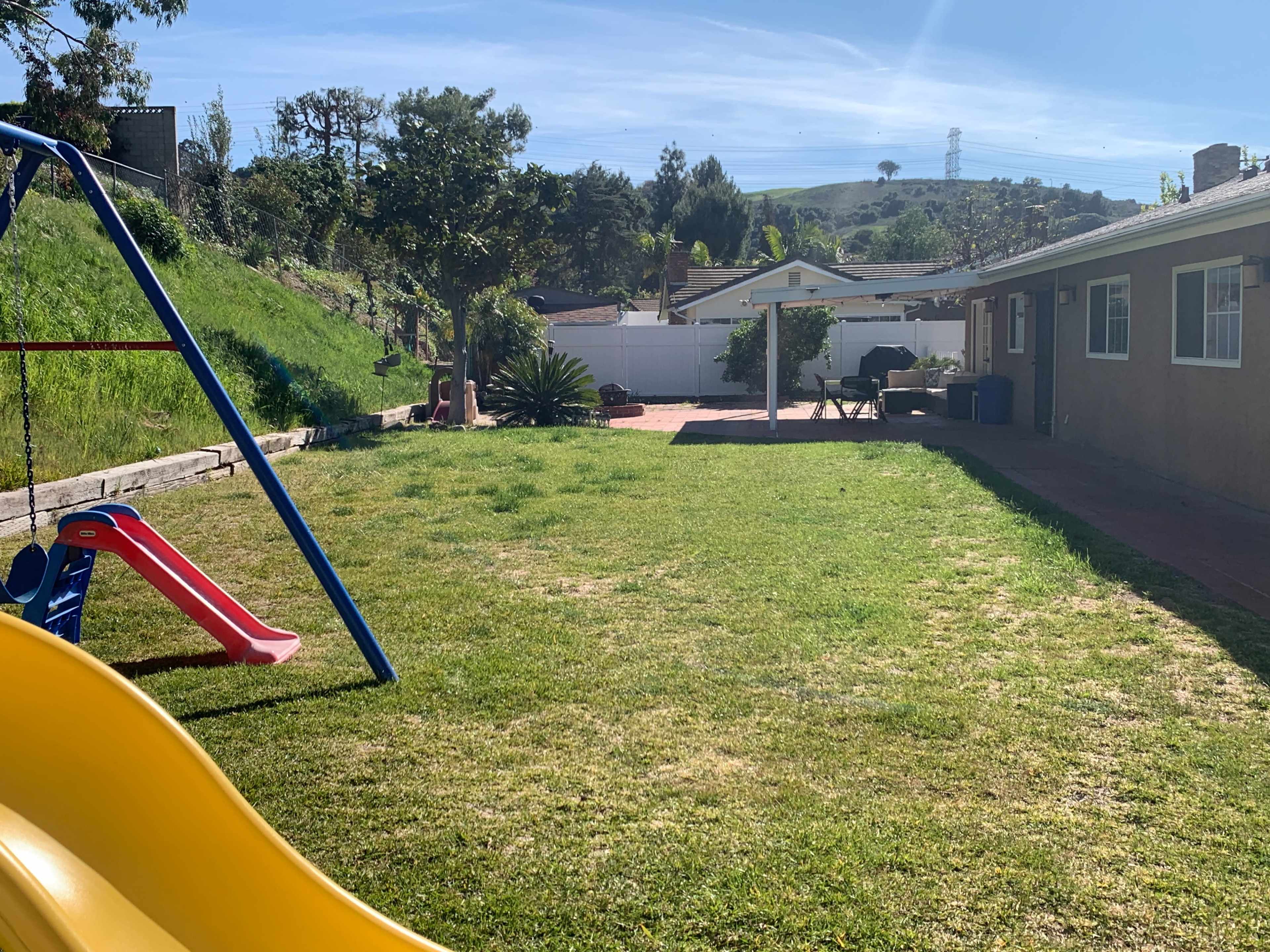 The image shows a spacious backyard with a swing set, a slide, and a patio area beside a house, set against a backdrop of trees and hills.