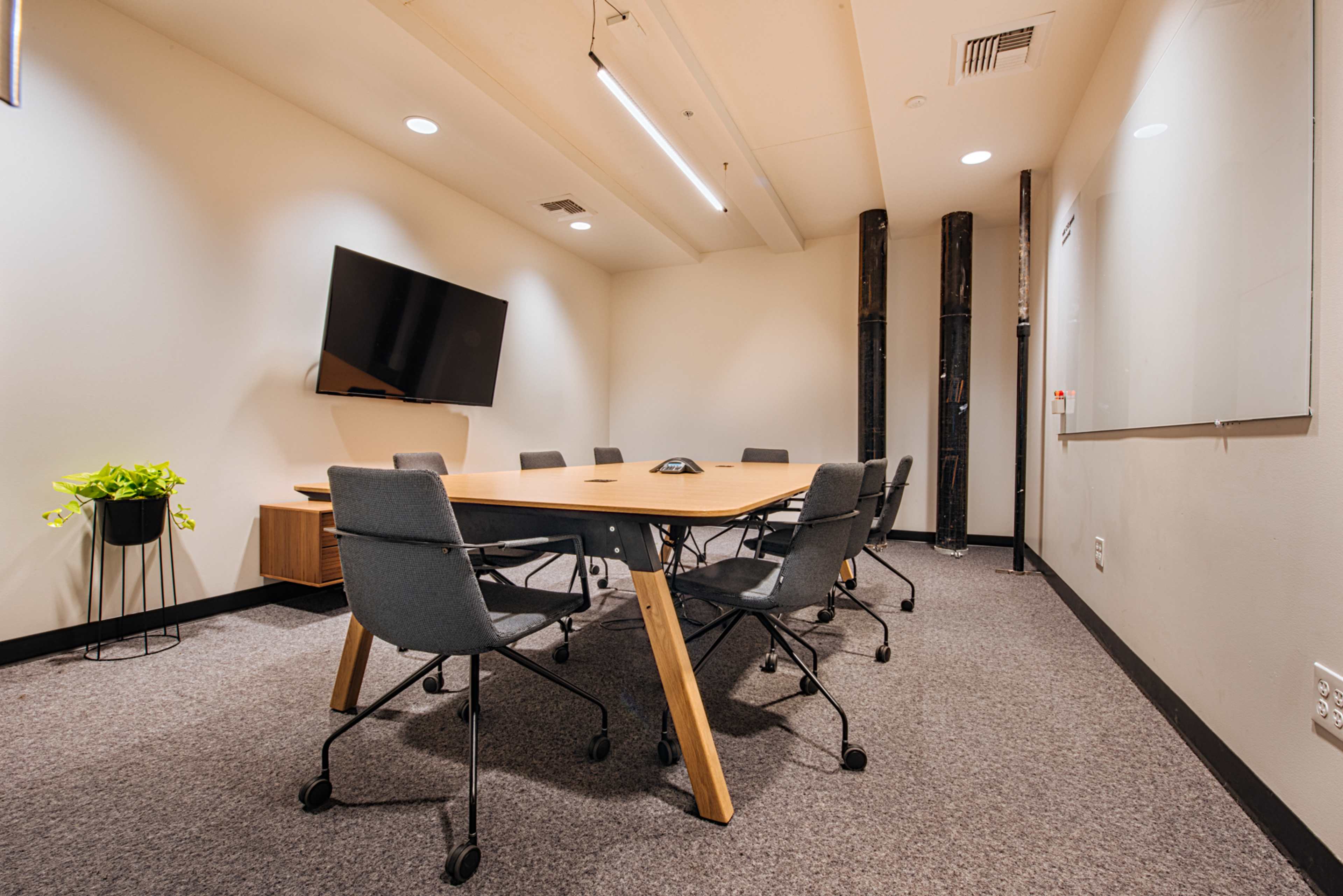 A conference room featuring a large table surrounded by chairs, a television on the wall, and a plant in a pot.