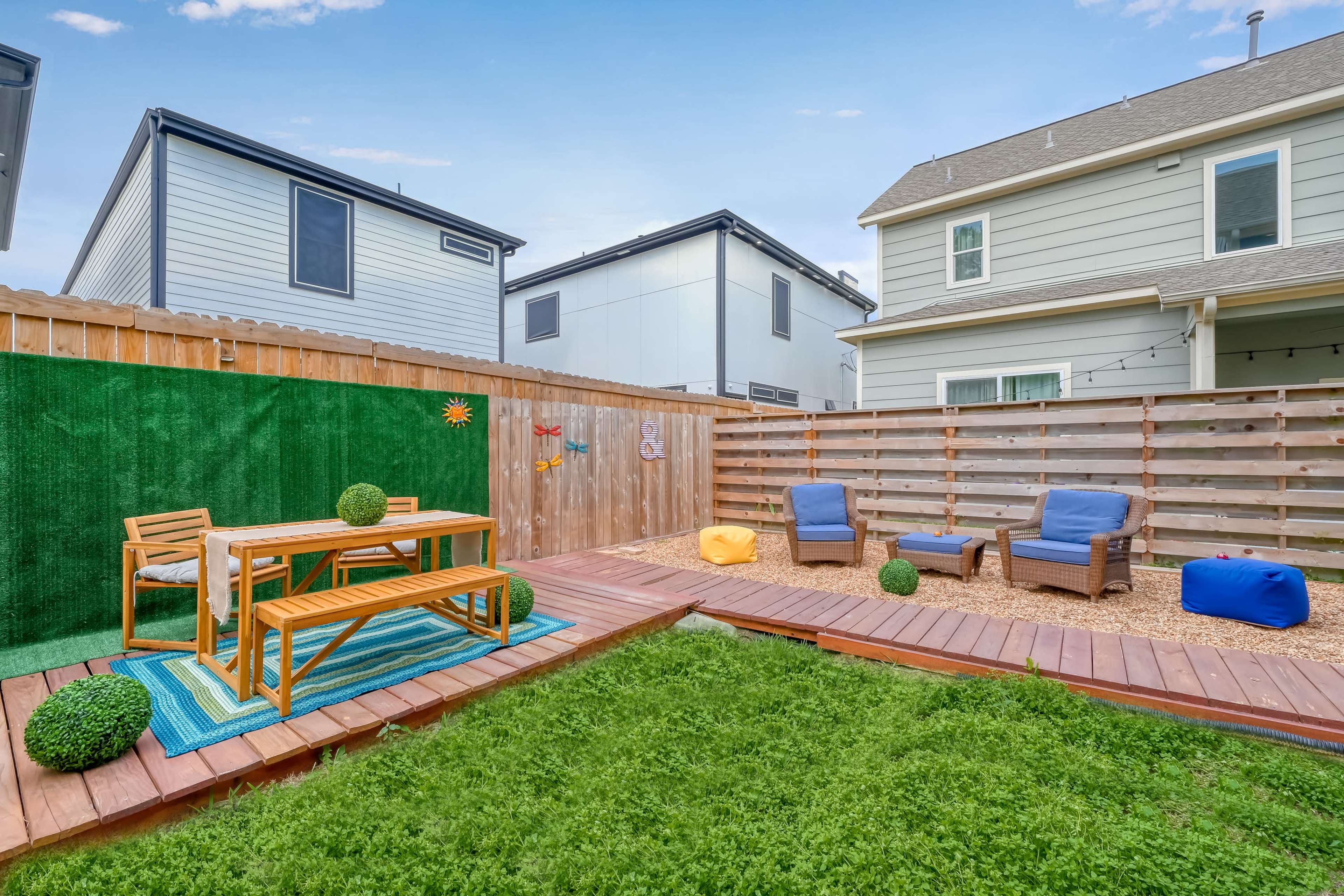 A backyard with a wooden deck, a green turf area, a table with chairs, and lounging chairs surrounded by wooden fencing.