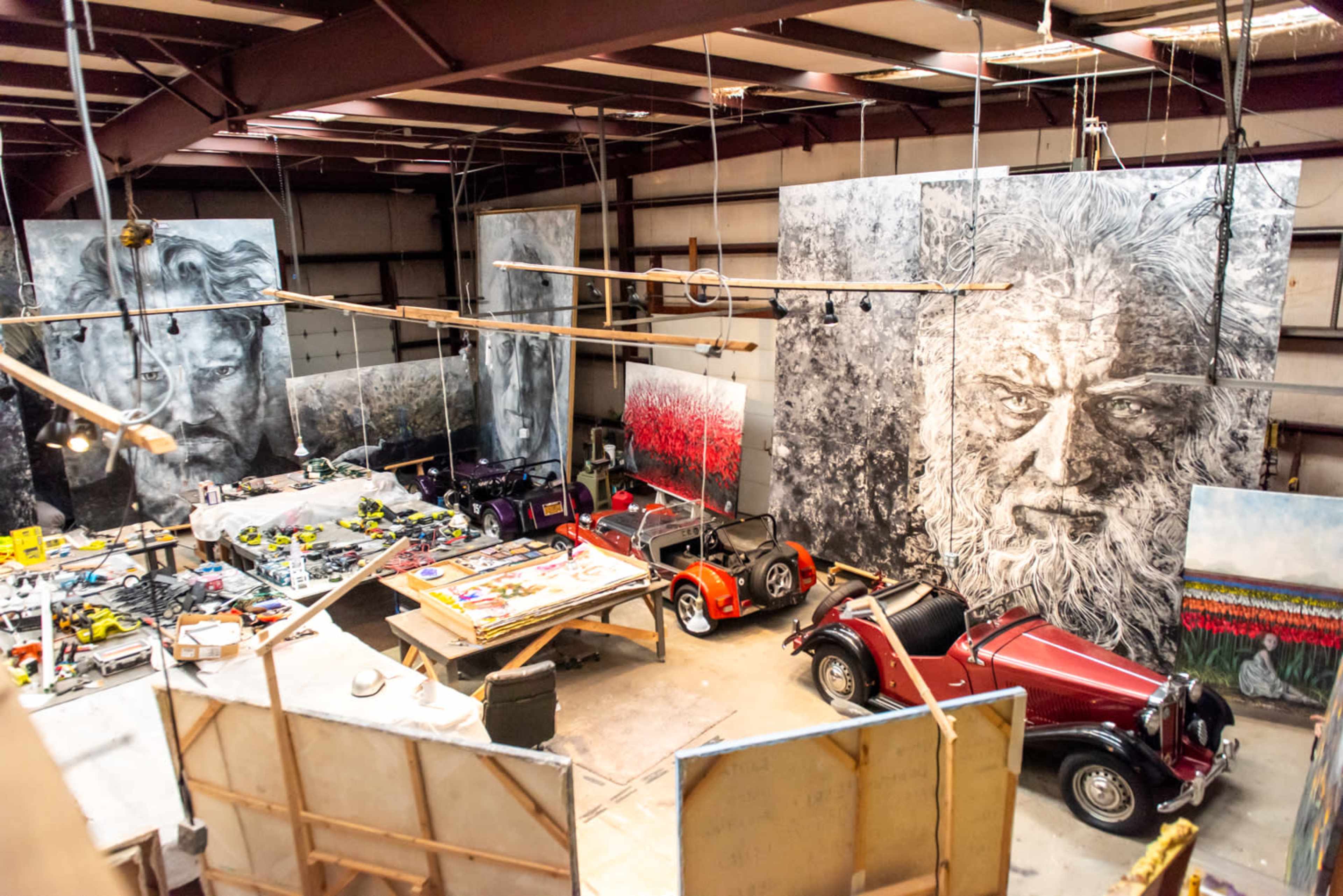 An expansive artist's studio filled with large, painted canvases and vintage cars amidst various art supplies and tools.
