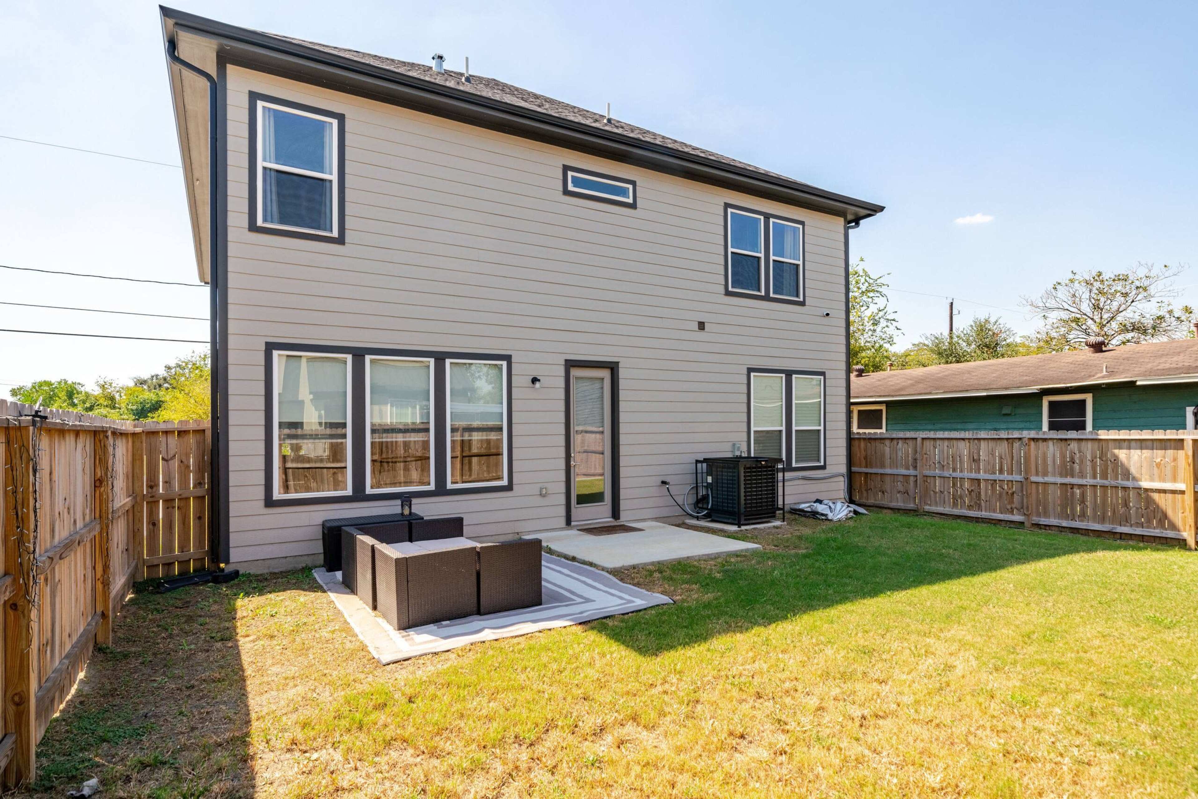 The image shows the back of a two-story house with a fenced yard, a concrete patio area, and outdoor furniture.