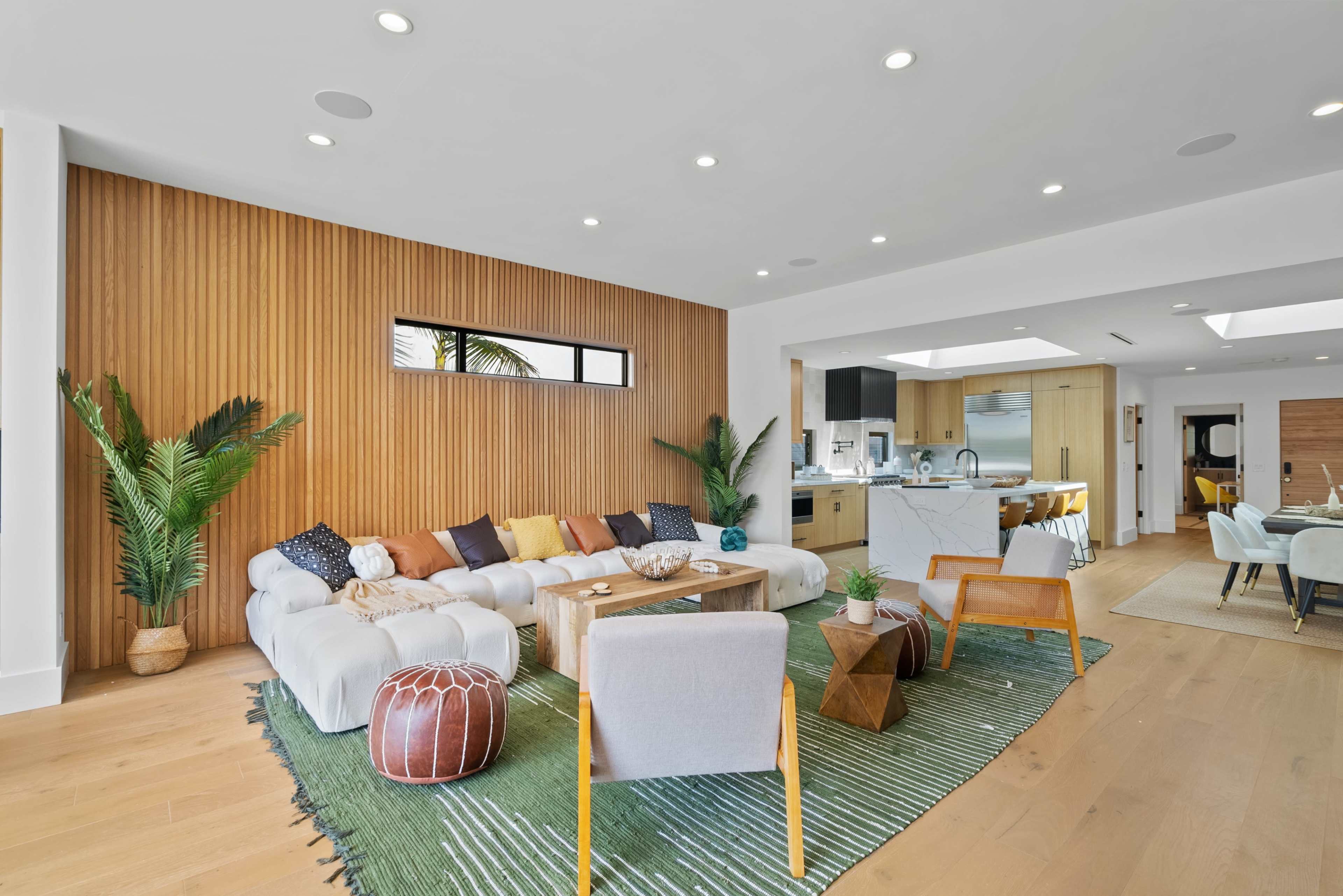 A modern living room with a sectional sofa, a wooden accent wall, a coffee table, and greenery, leading to an open kitchen and dining area.