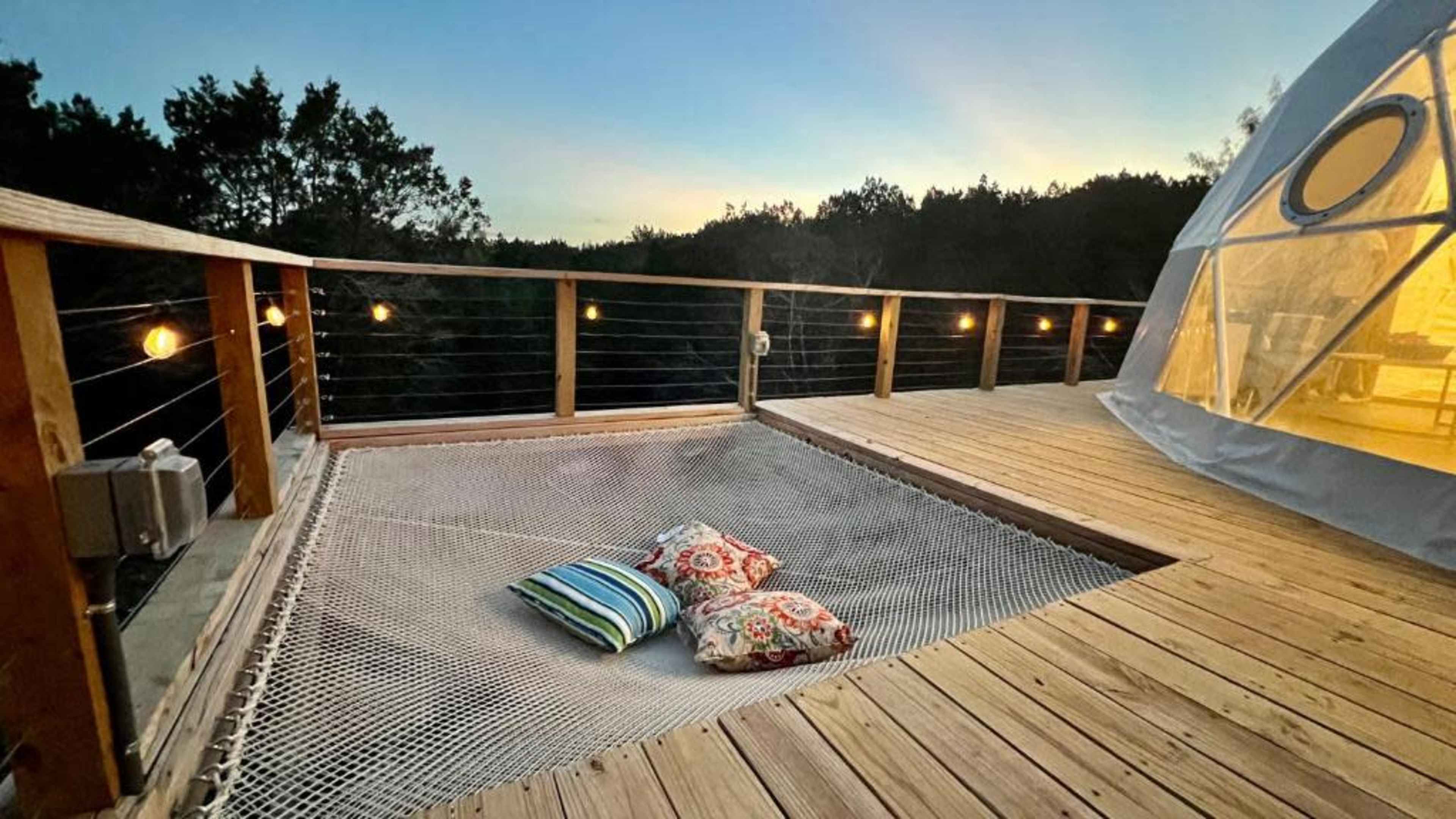 A wooden deck features a netted seating area with colorful pillows and is surrounded by trees at twilight.