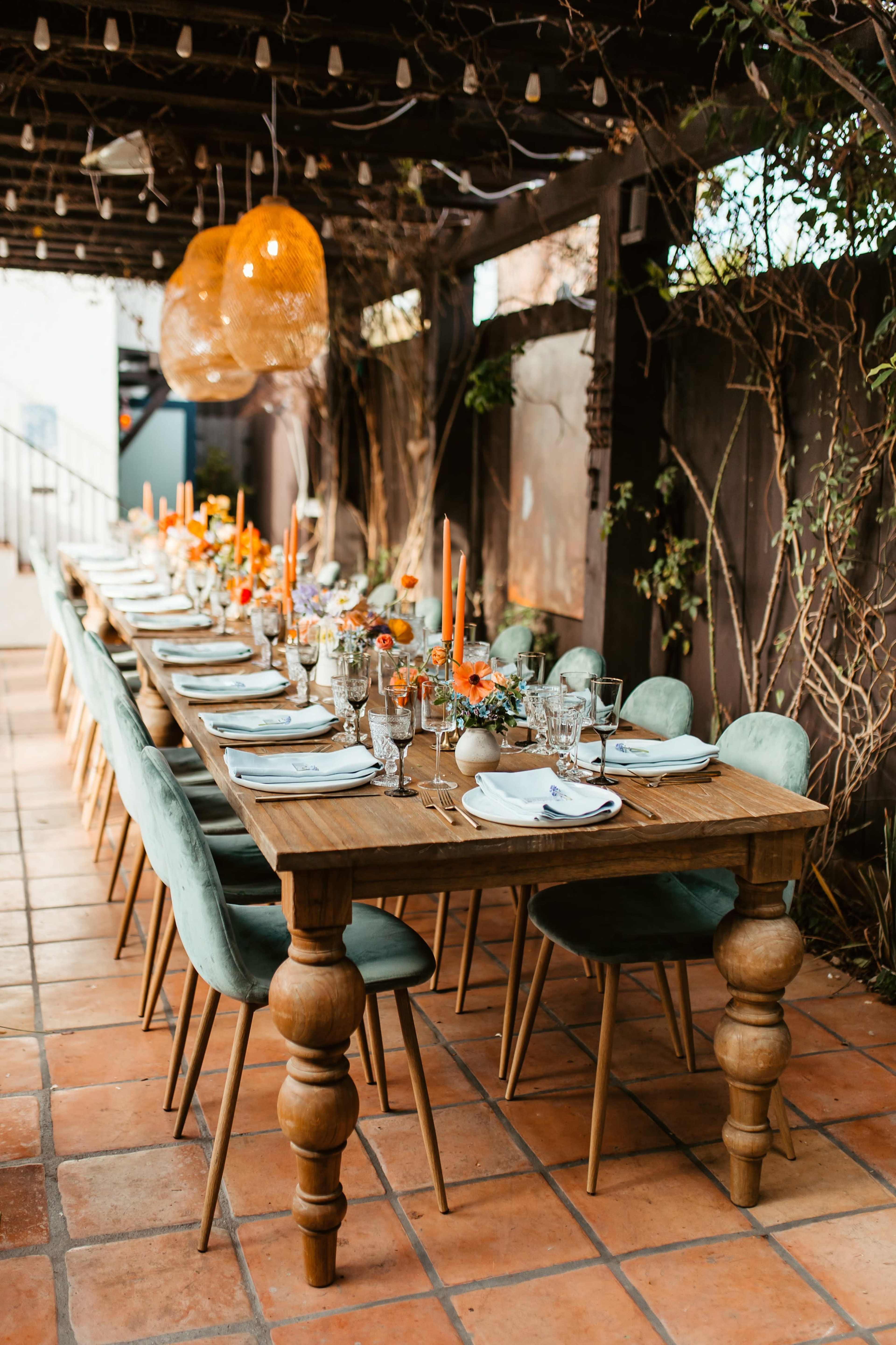A long wooden dining table is elegantly set with plates, glasses, and orange floral arrangements under hanging lights in a garden-like setting.