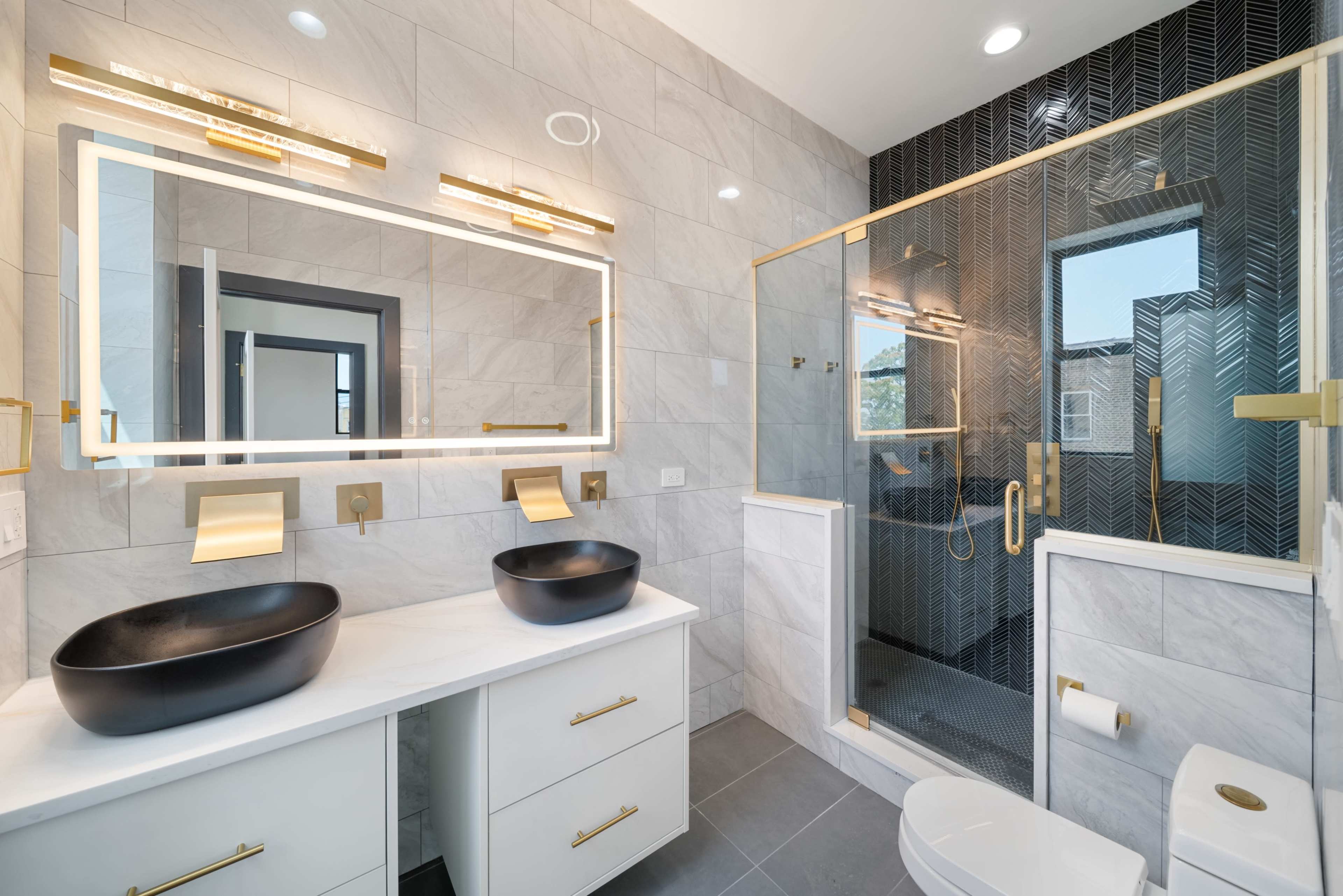 The image shows a modern bathroom featuring a double vanity with black vessel sinks, gold fixtures, a large illuminated mirror, and a glass shower with a herringbone tile design.