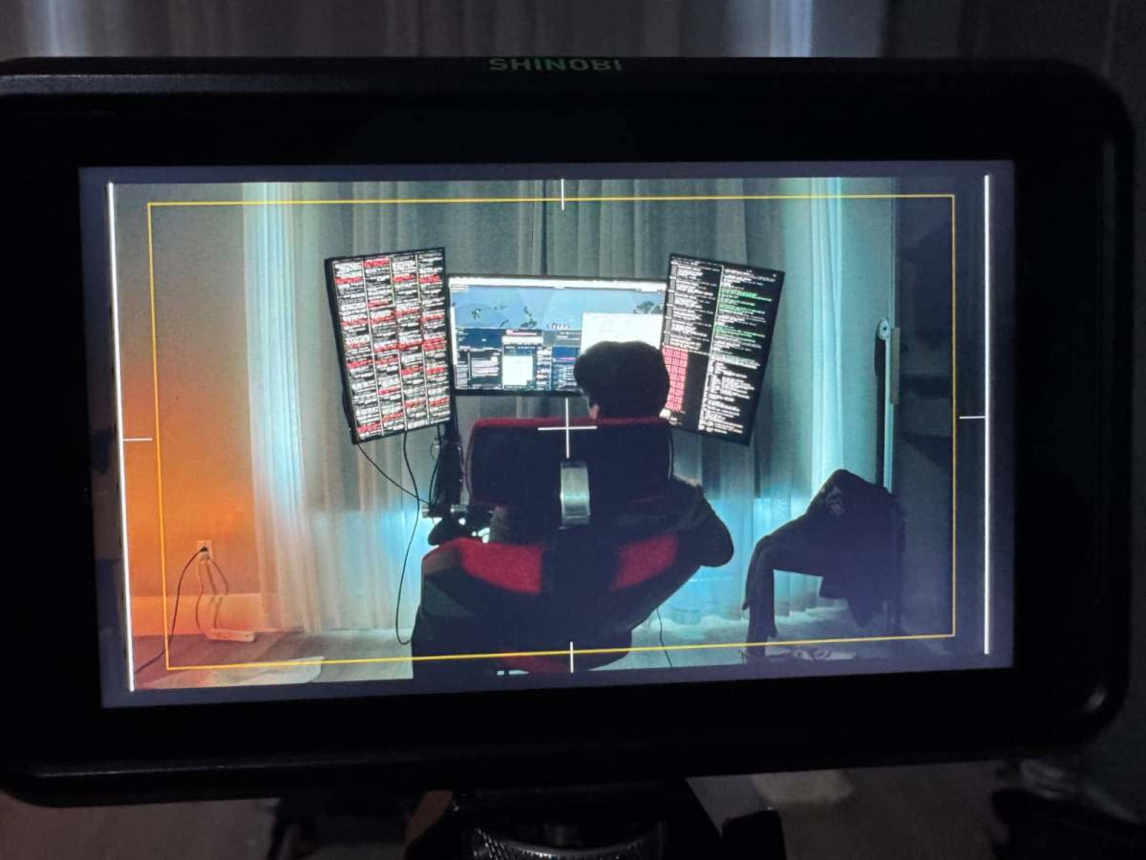 A person sits in a gaming chair facing multiple computer monitors displaying data and graphics in a dimly lit room.
