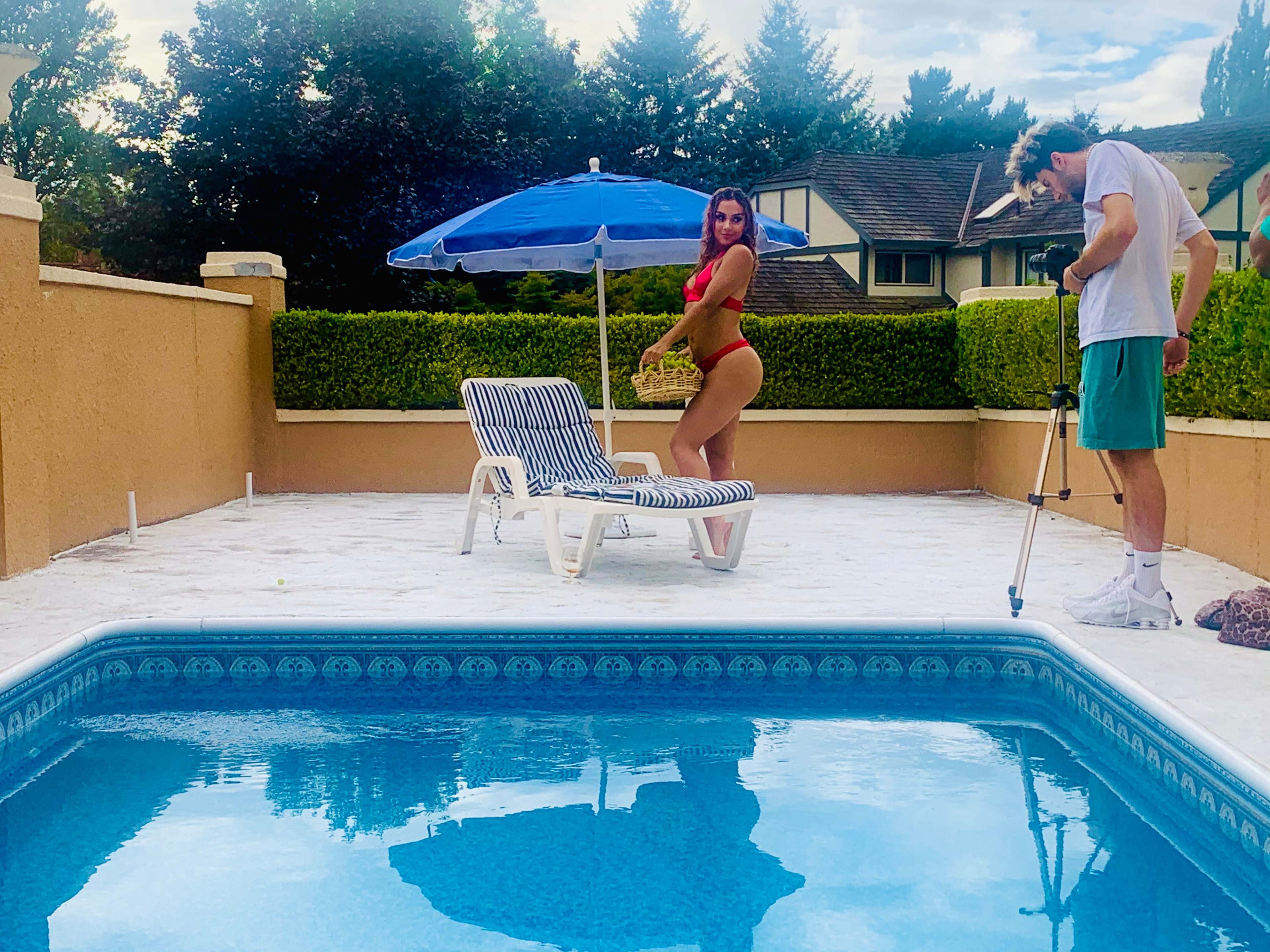 A woman in a red swimsuit poses by a swimming pool while a man adjusts a camera on a tripod nearby.