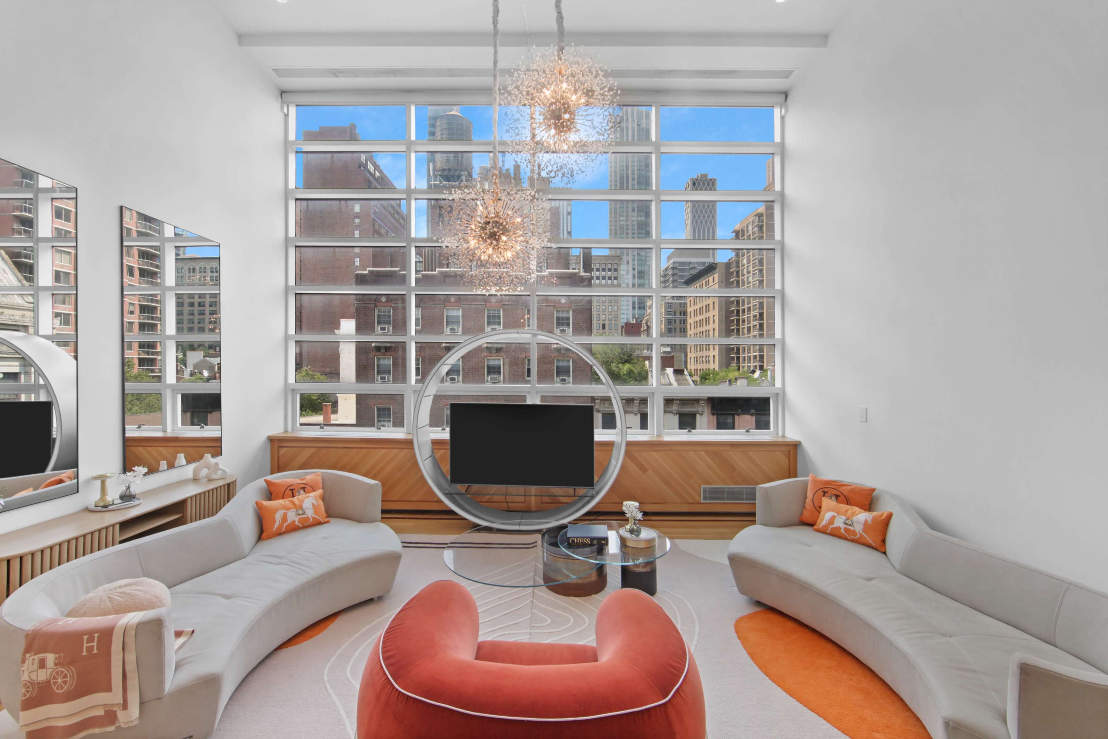 The spacious living room features large windows that showcase a city view, with modern furniture arranged around a central television.