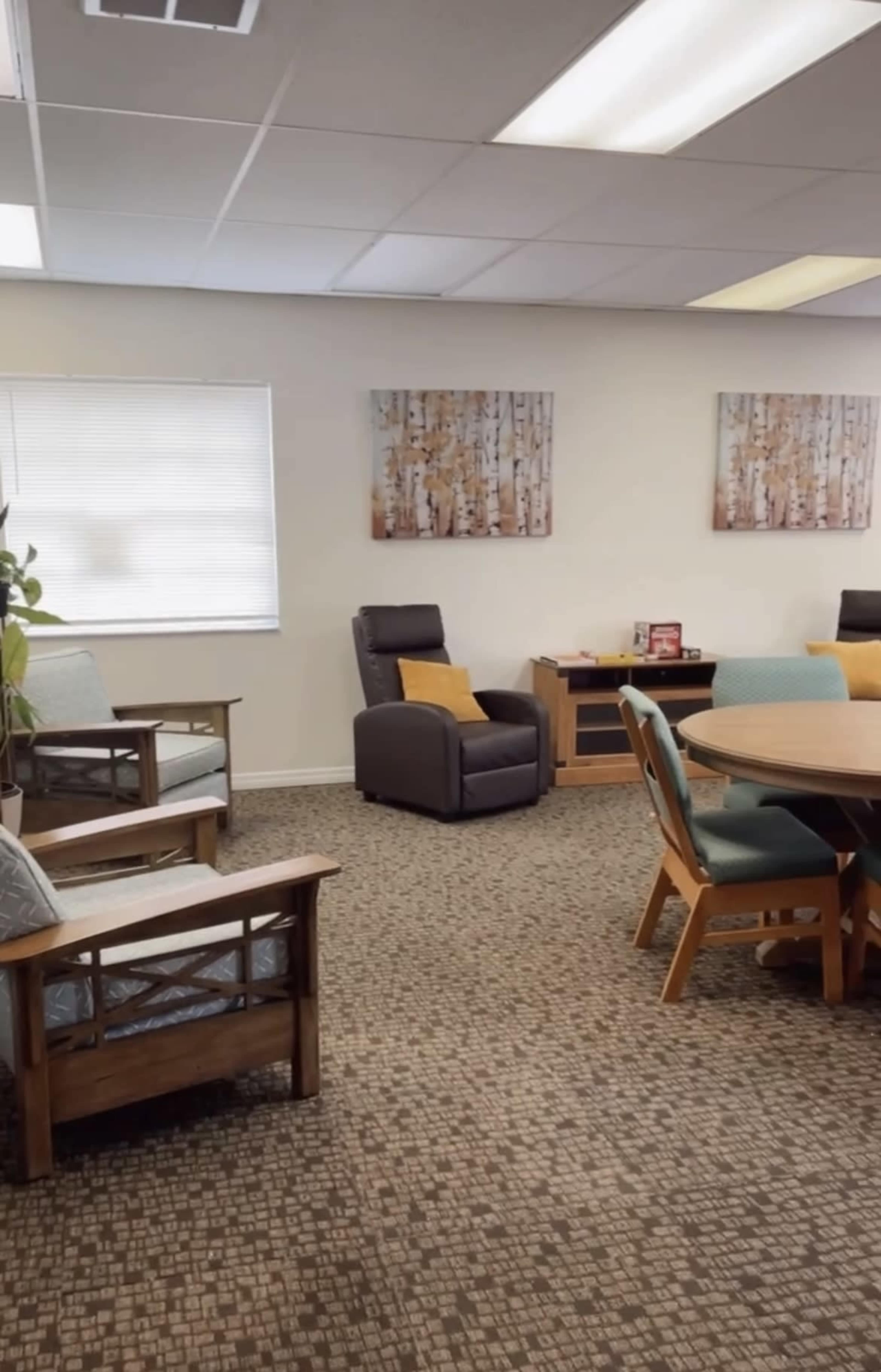 A room with two wooden chairs, a reclining chair, a round table with wooden chairs, and wall art depicting trees.