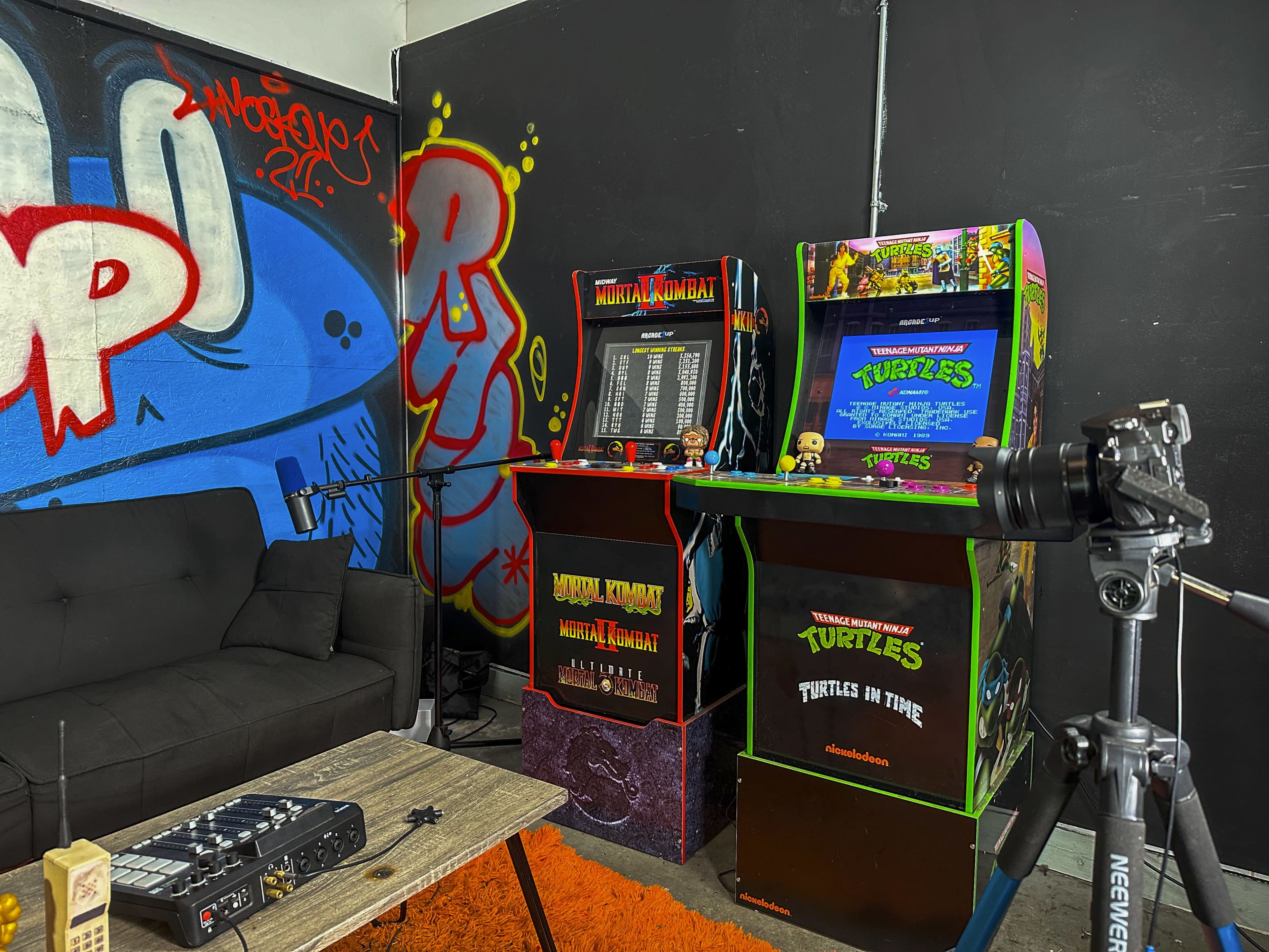 A corner of a room featuring two vintage arcade machines, one for "Mortal Kombat" and the other for "Teenage Mutant Ninja Turtles," alongside a black sofa and a table with audio equipment on it.
