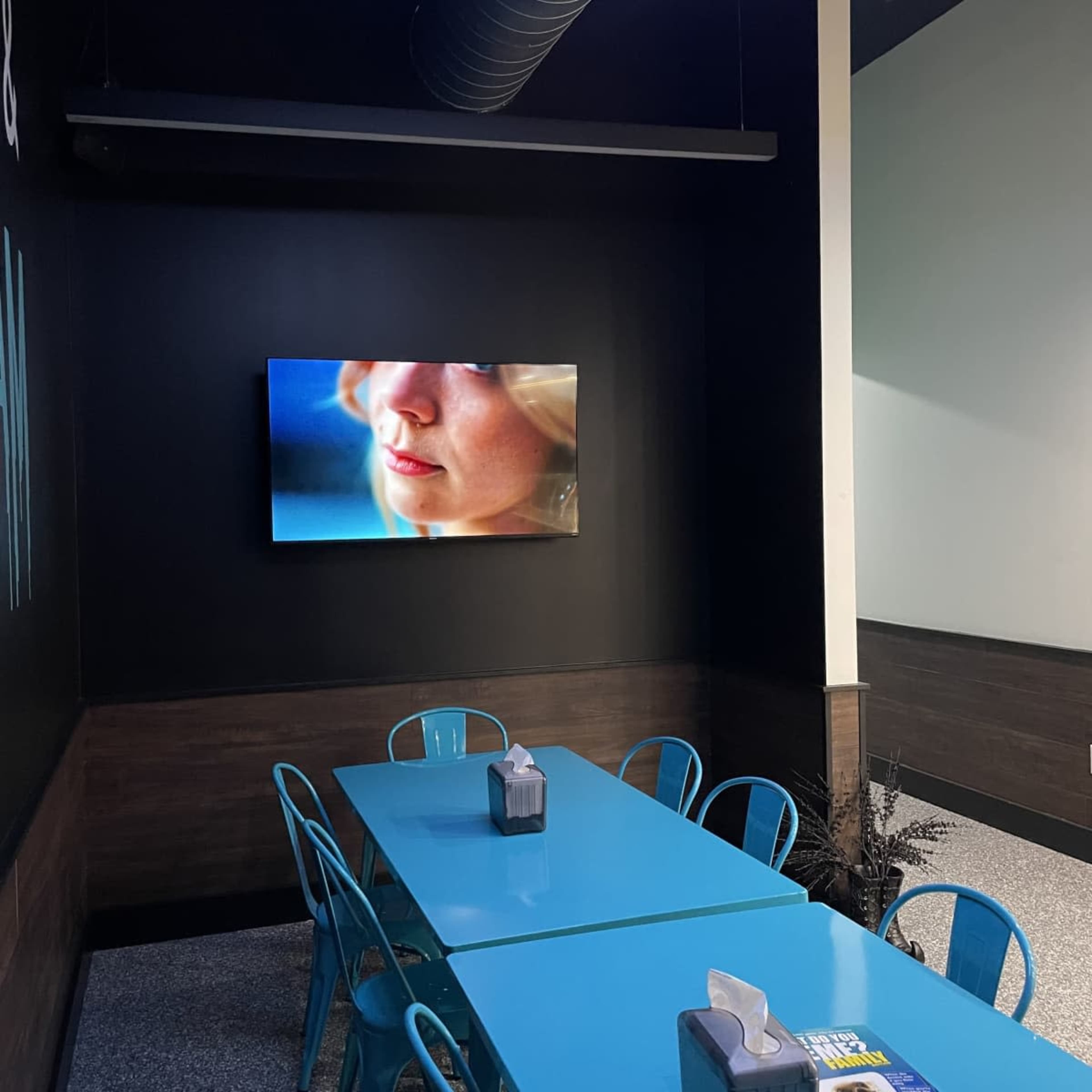 A brightly lit dining area features a blue table surrounded by metal chairs, with a television mounted on the black wall displaying an image of a person's face.