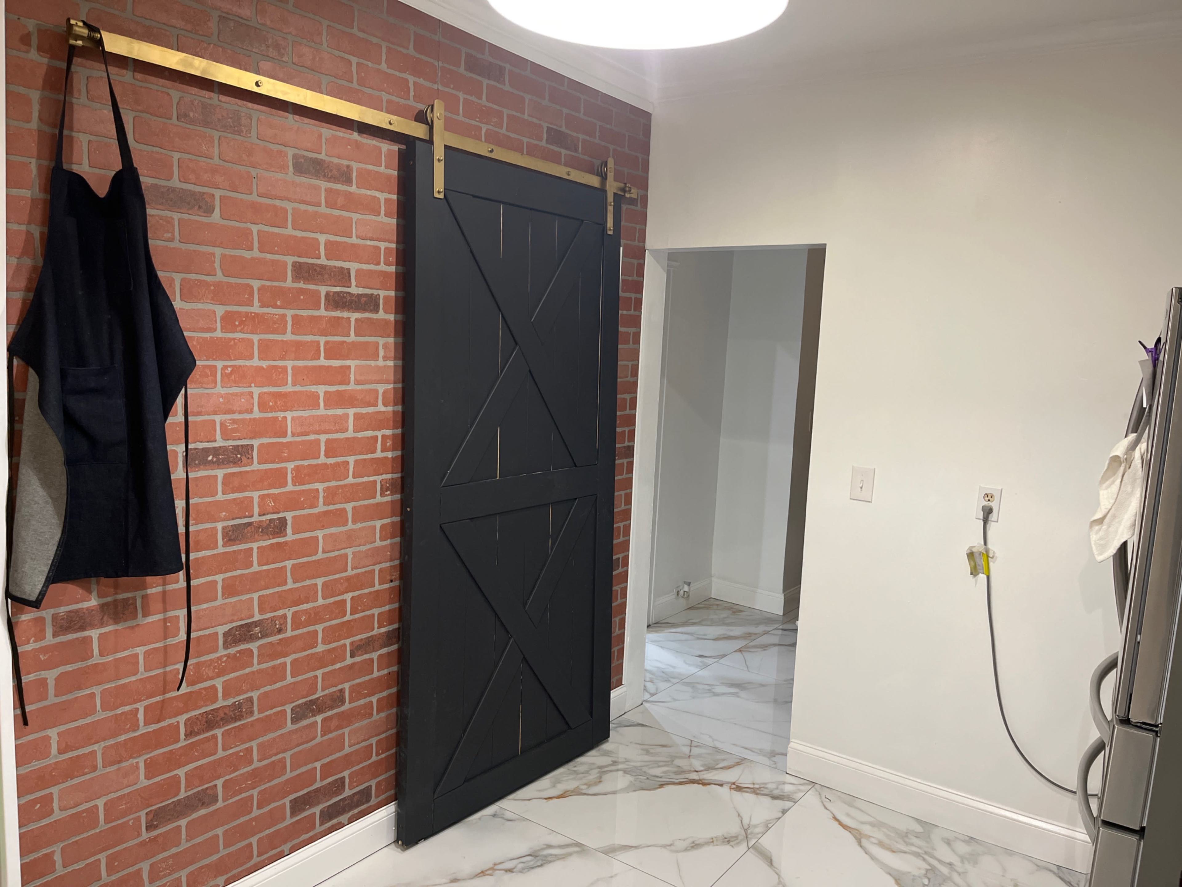 The image shows a kitchen area featuring a black sliding barn door on a brick wall, with an apron hanging beside it and white marble flooring.