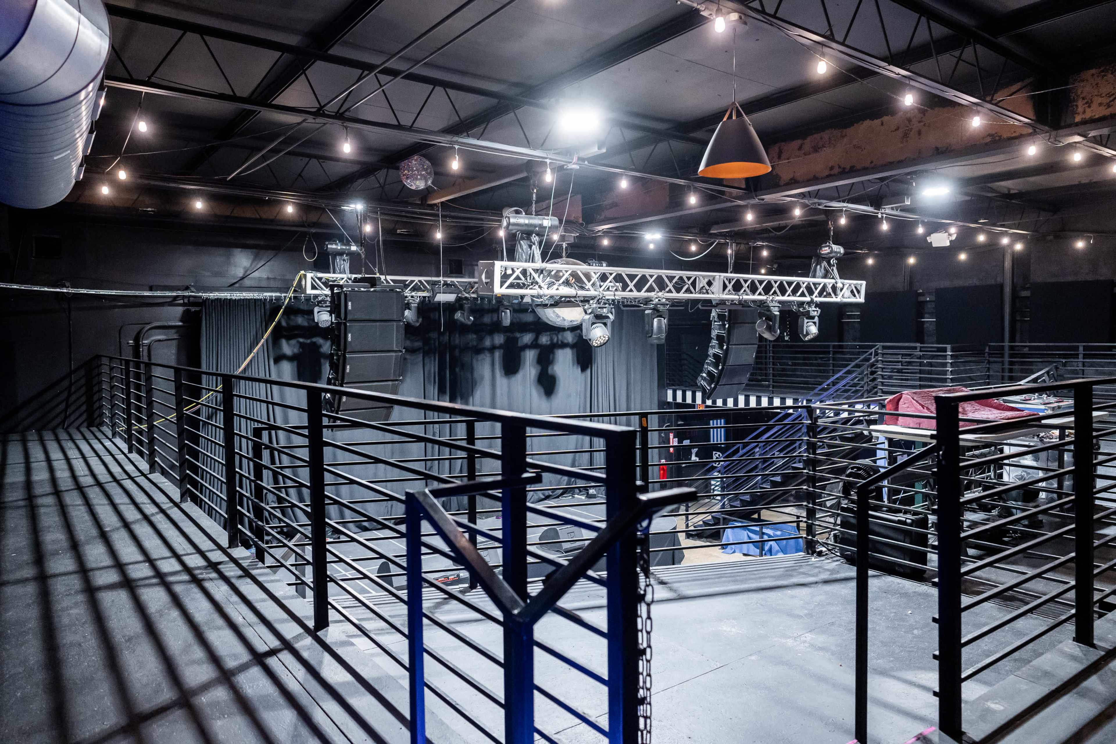 An empty indoor venue with metal railings, hanging lights, and suspended sound equipment.