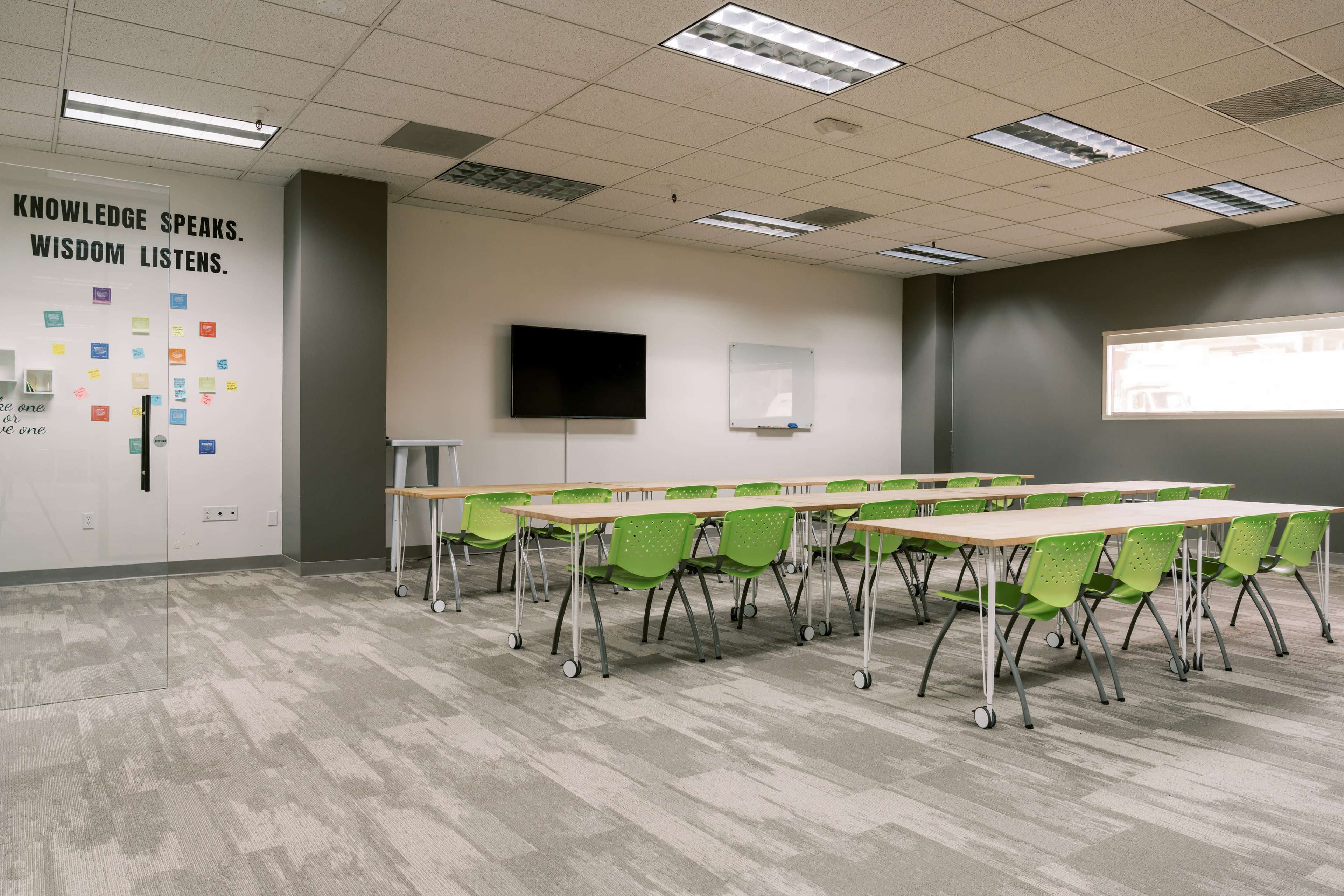The image shows a classroom with green chairs and tables arranged in rows, featuring a wall with a quote and a television screen.