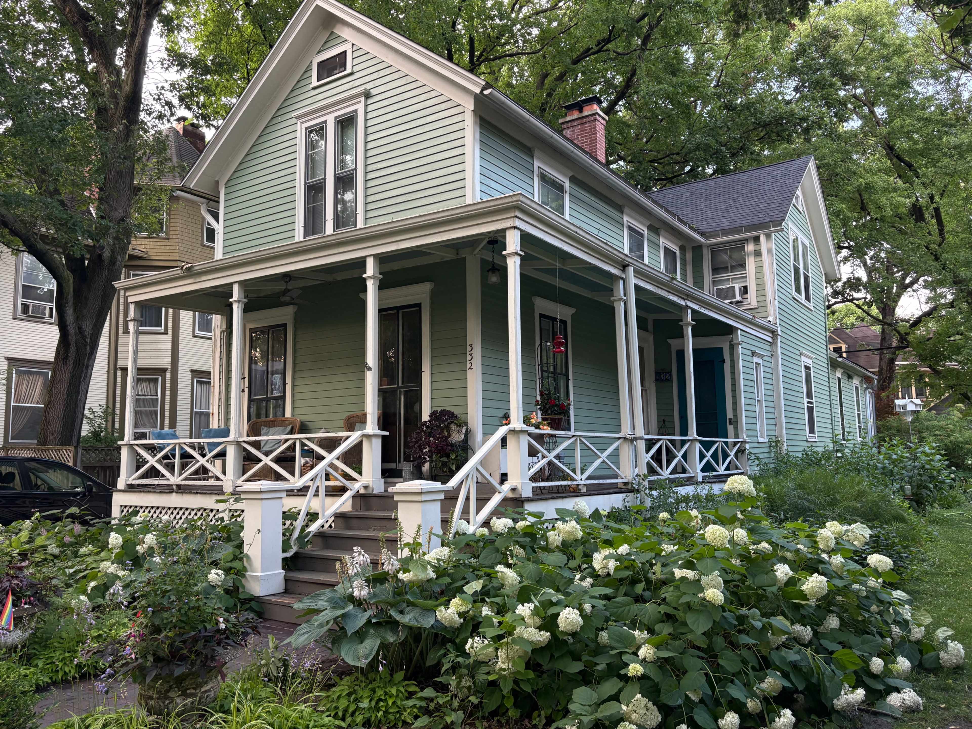 The image shows a light blue house with a large porch, set in a landscaped area with greenery and flowering plants.