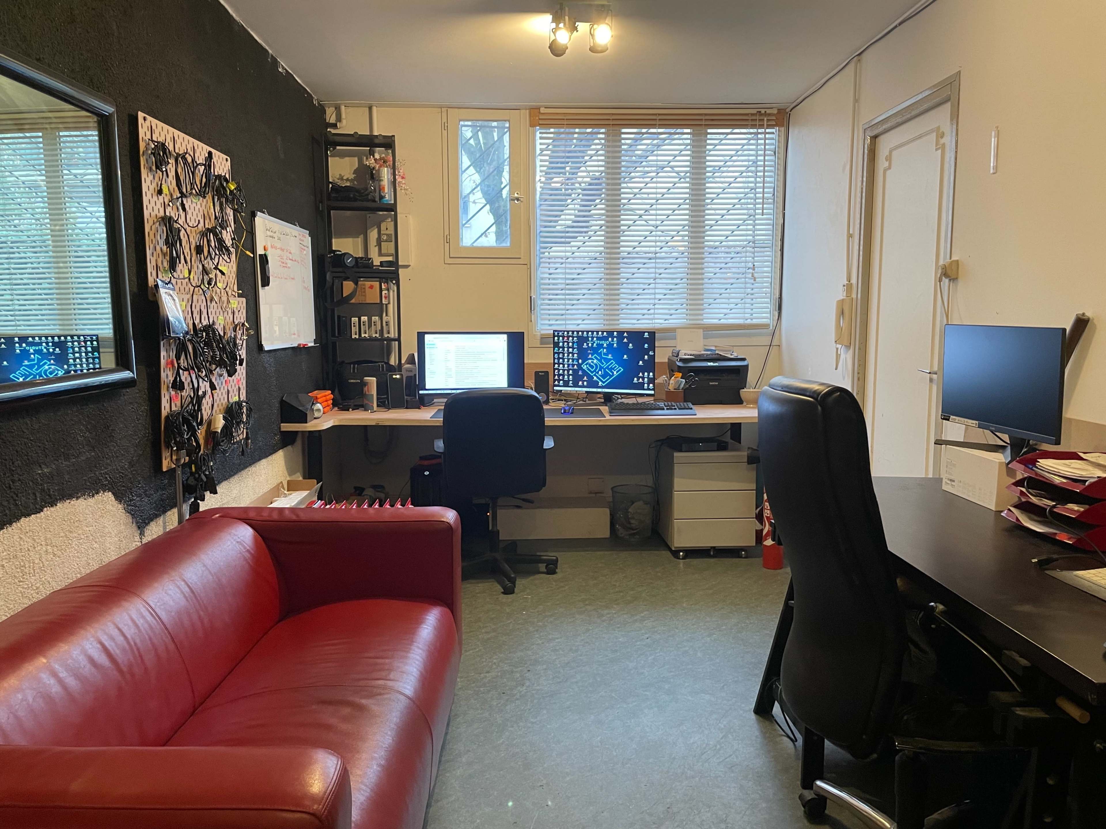 An office space with a red couch, two computer desks with monitors, and a large window providing natural light.