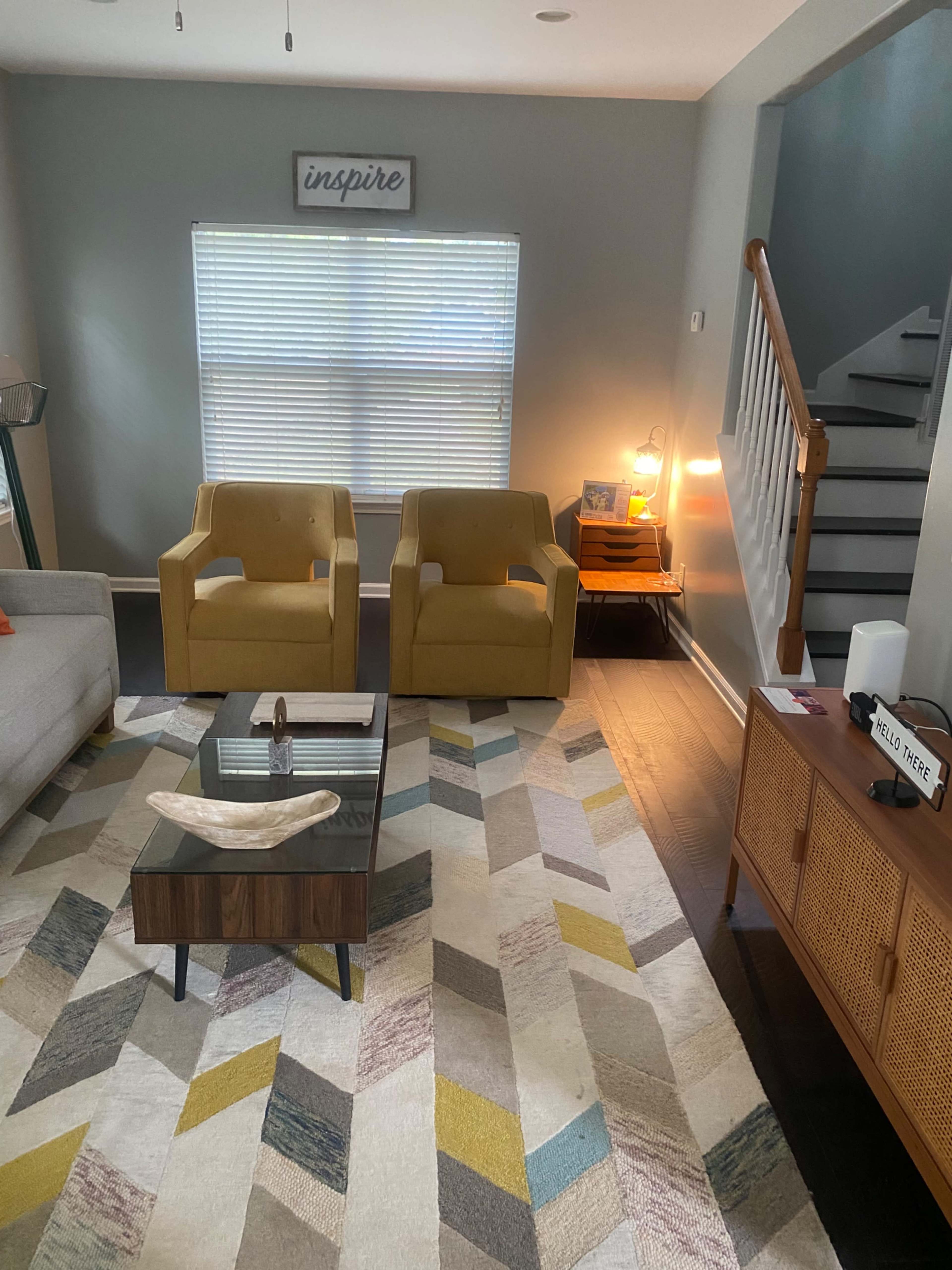 The living room features two yellow armchairs facing a coffee table on a multicolored geometric rug, with a sofa and a staircase visible in the background.