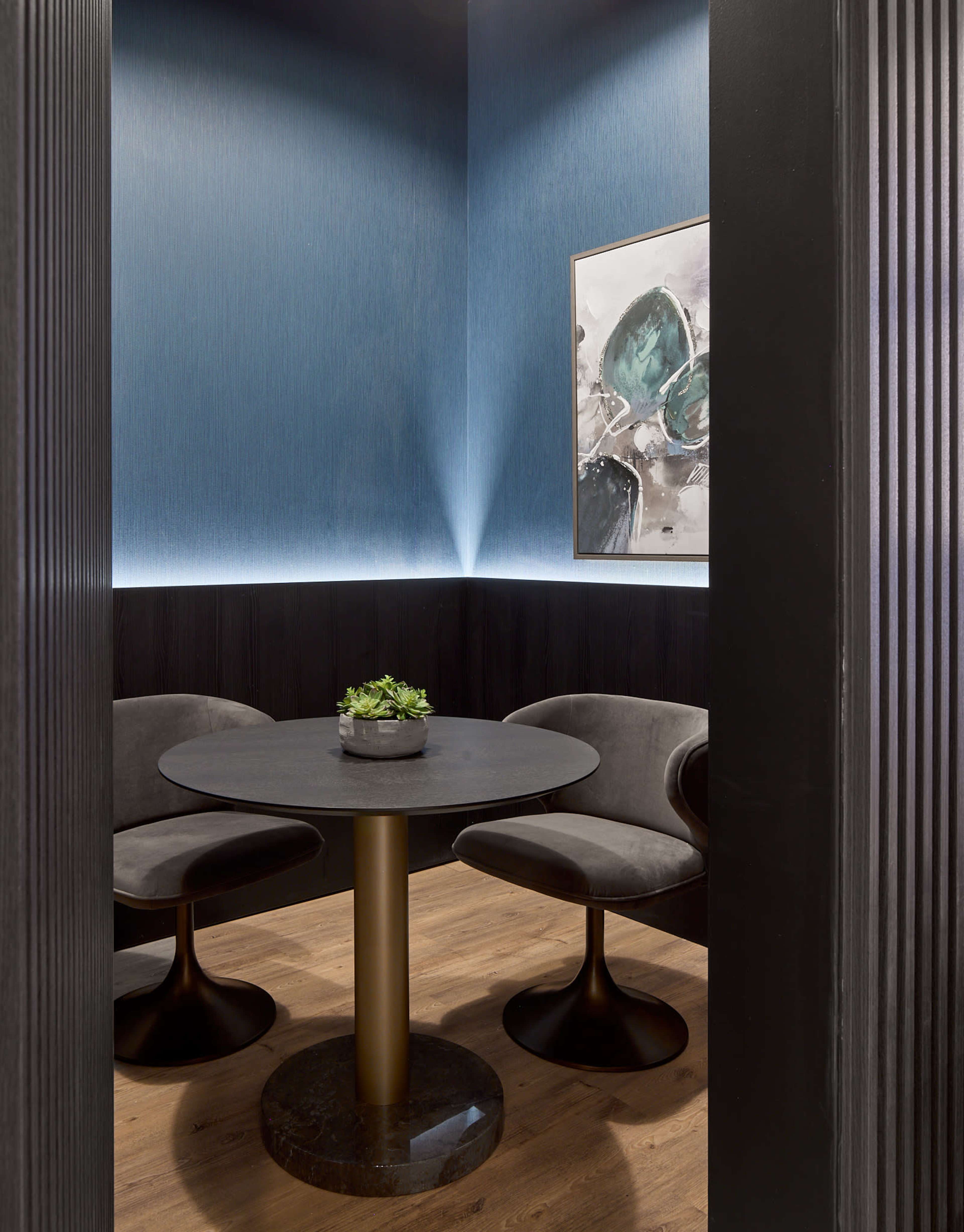 A small, modern dining area features a round black table with two gray chairs, surrounded by blue walls and illuminated by soft lighting.