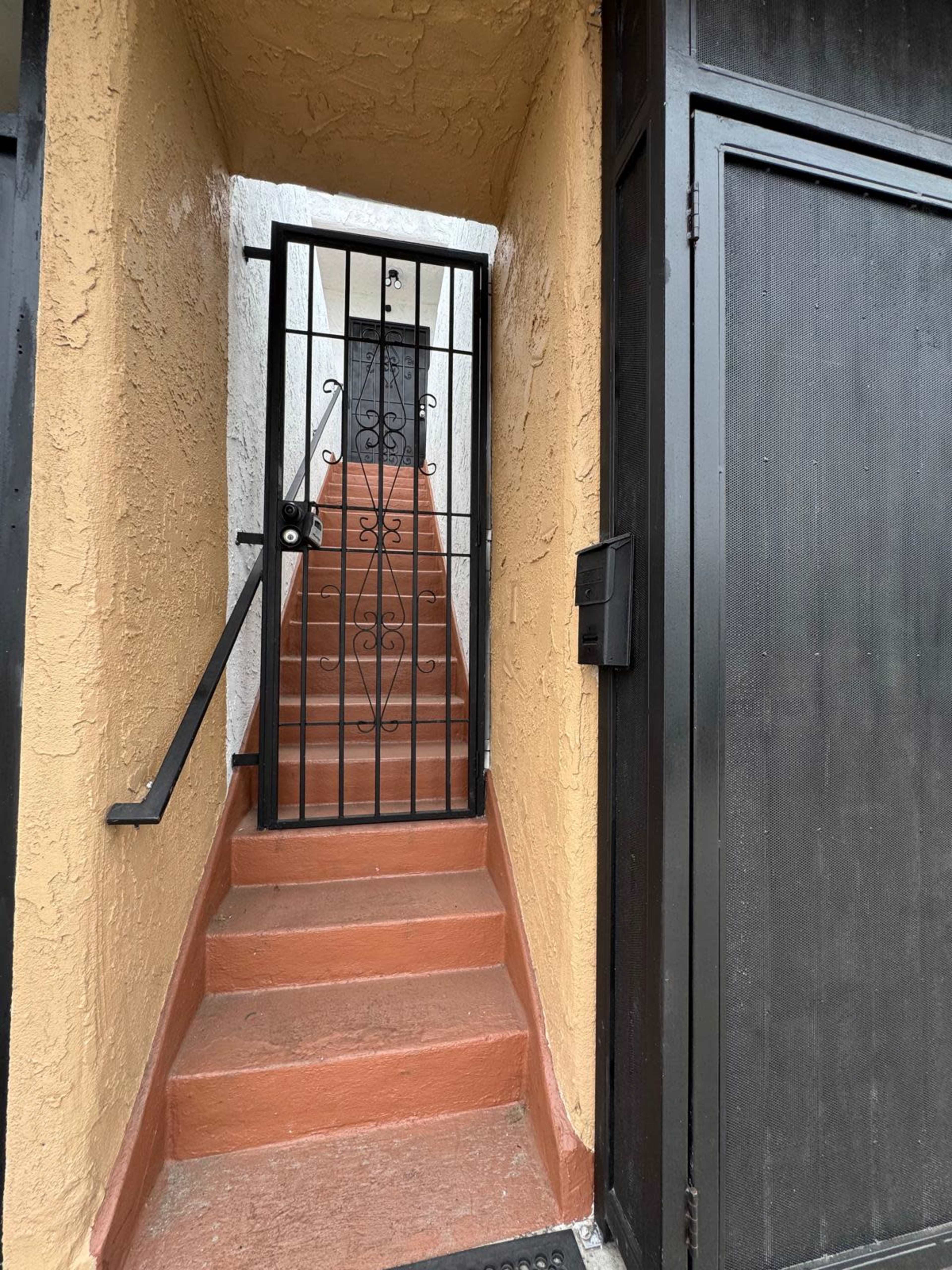 The image shows a narrow staircase leading up to a gated entrance, flanked by an orange wall and black gate.