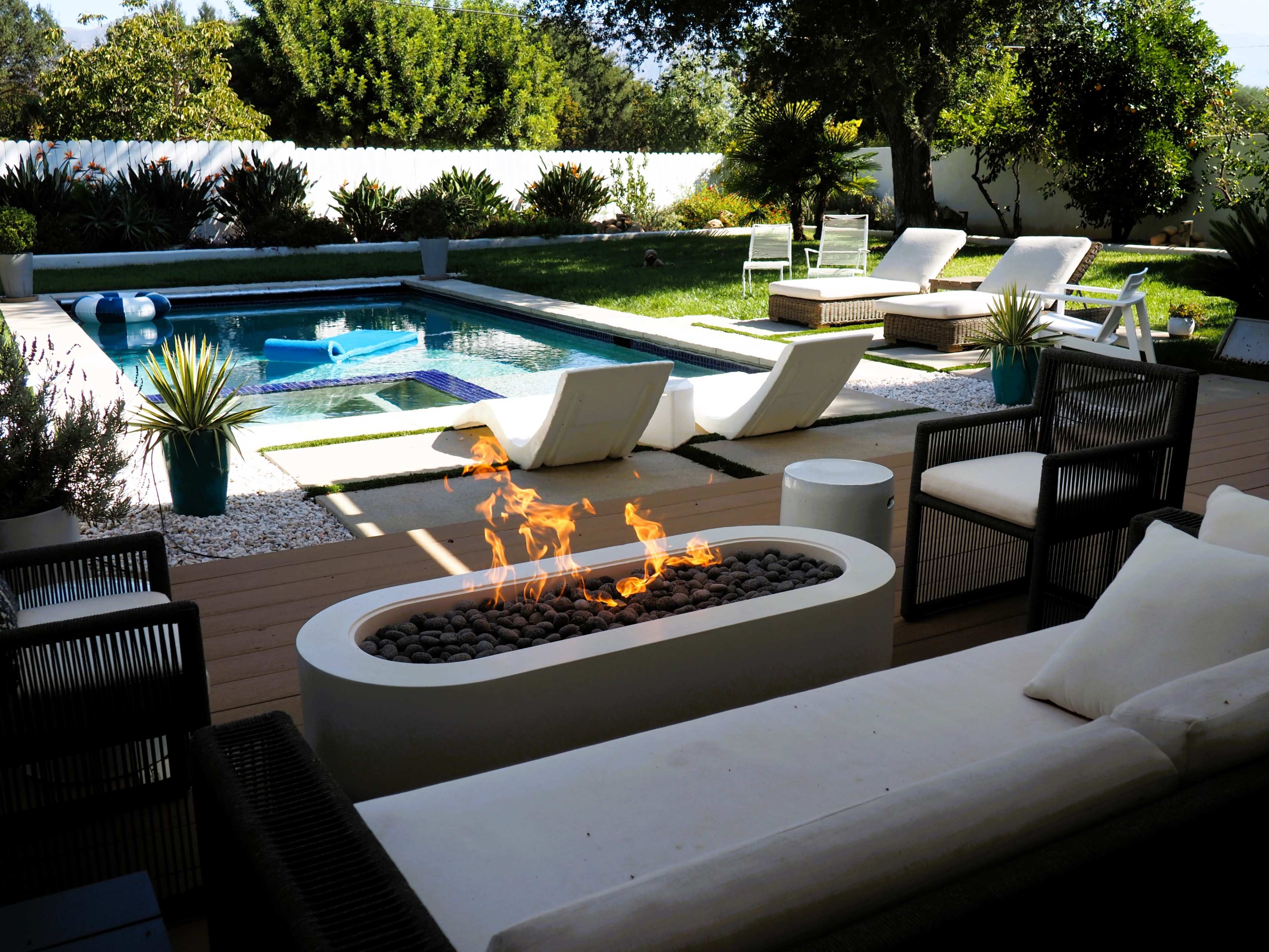 The image shows a modern outdoor patio with a pool, lounge chairs, and a fire feature surrounded by landscaping.