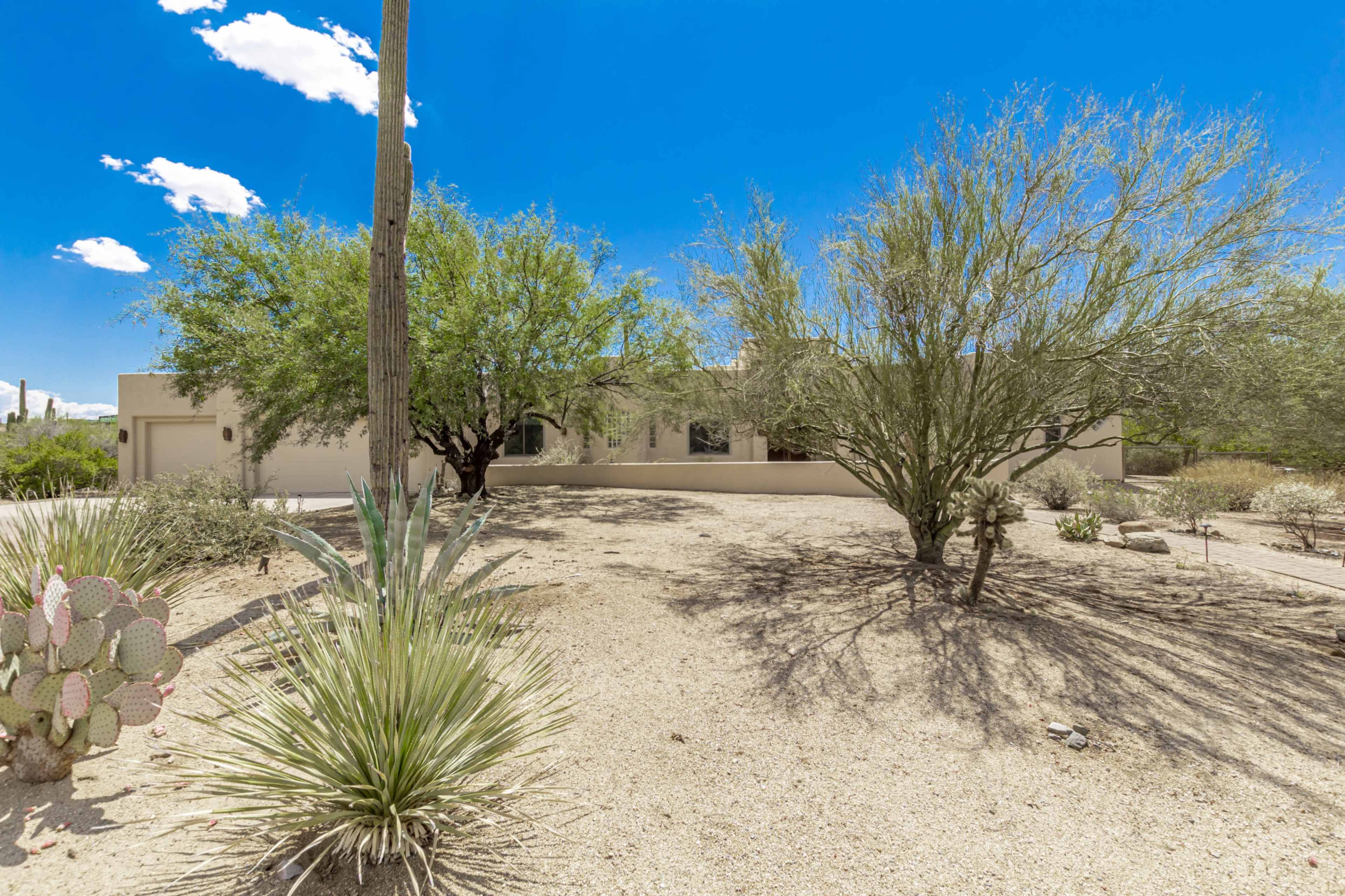 A modern home is set against a clear blue sky, surrounded by desert landscaping featuring cacti and low shrubs.