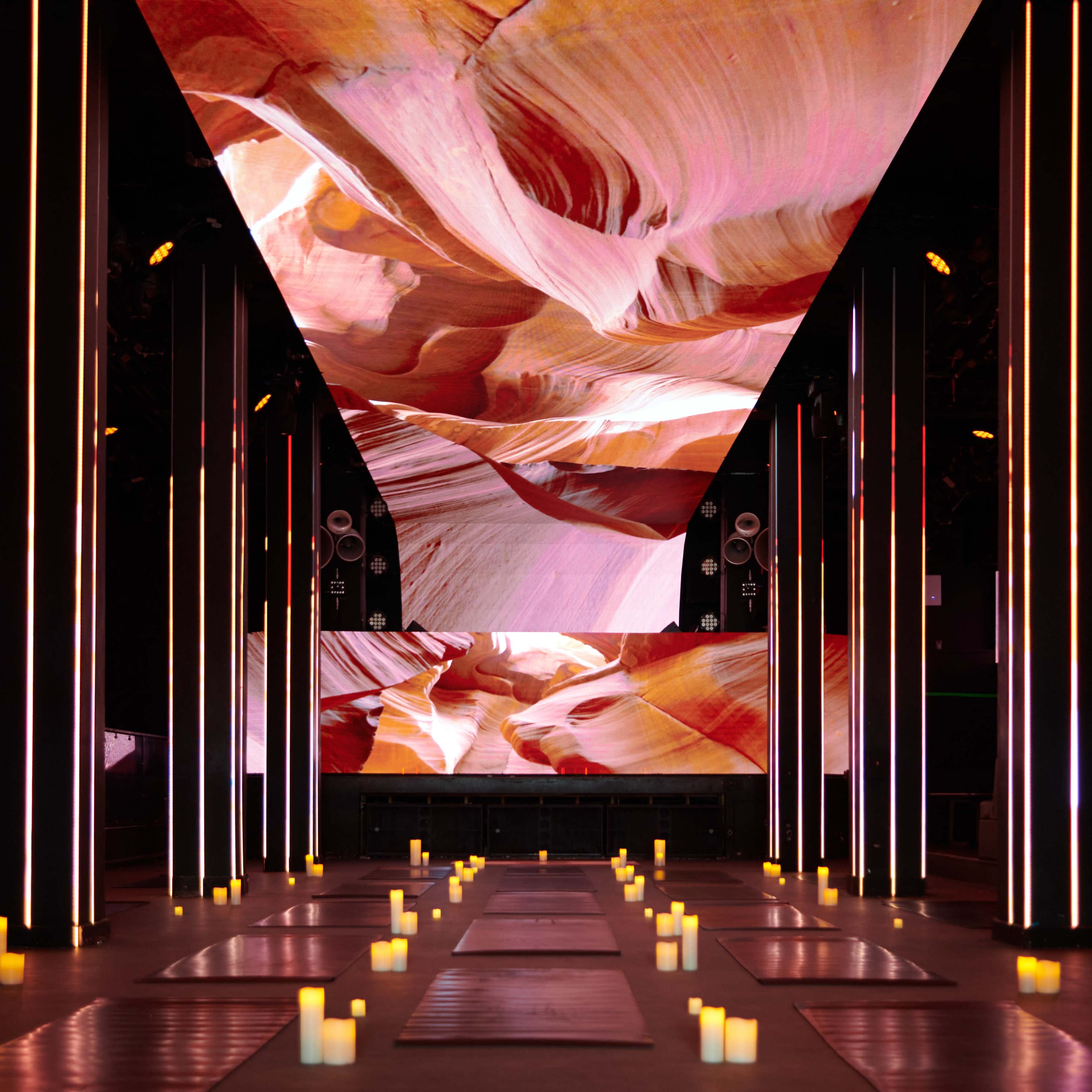 A dimly lit venue with illuminated candles lining the floor and a large digital display featuring colorful rock formations on the ceiling.