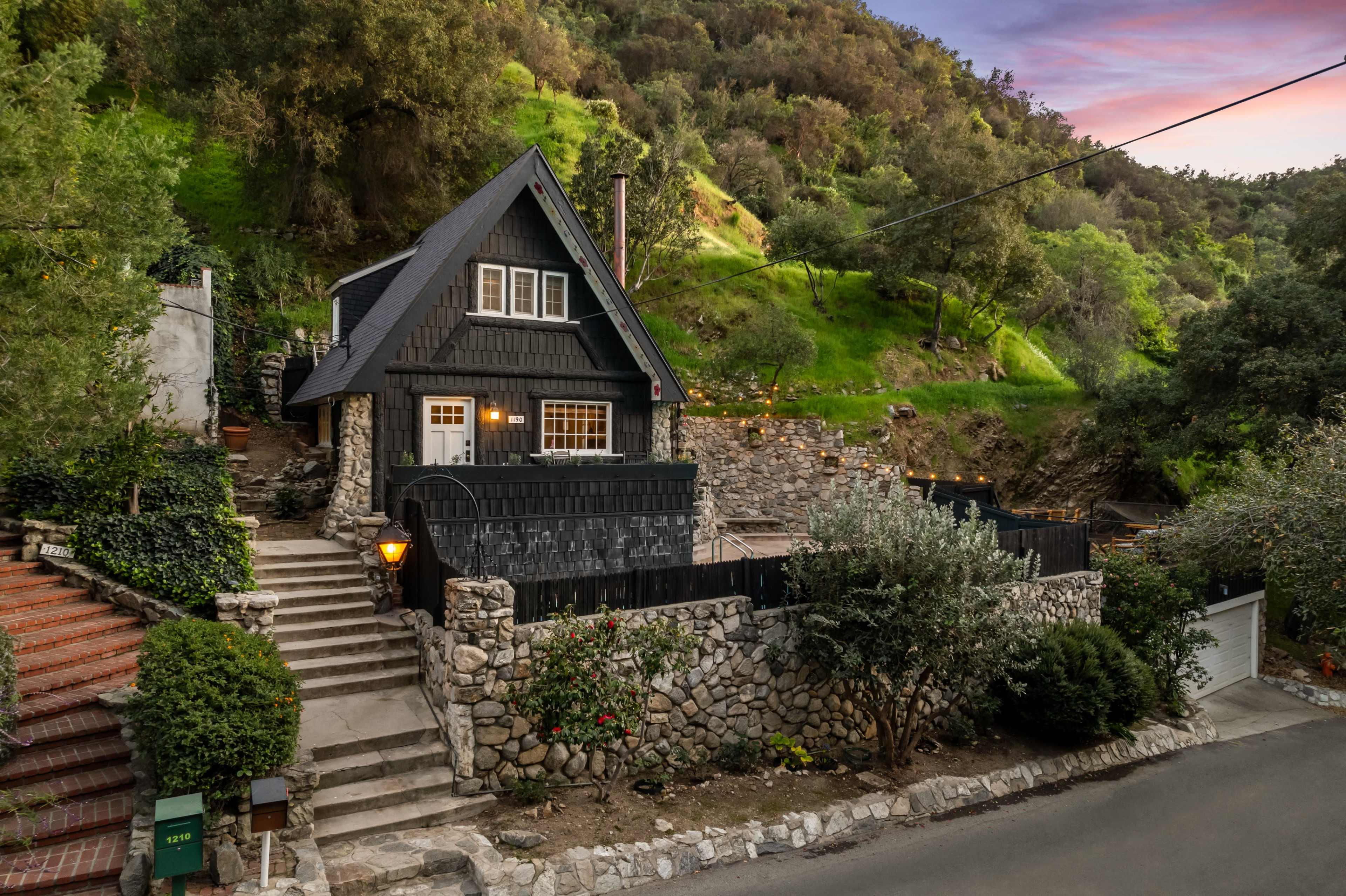 A charming house with a steeply pitched roof is situated on a hillside, surrounded by greenery and stone pathways.