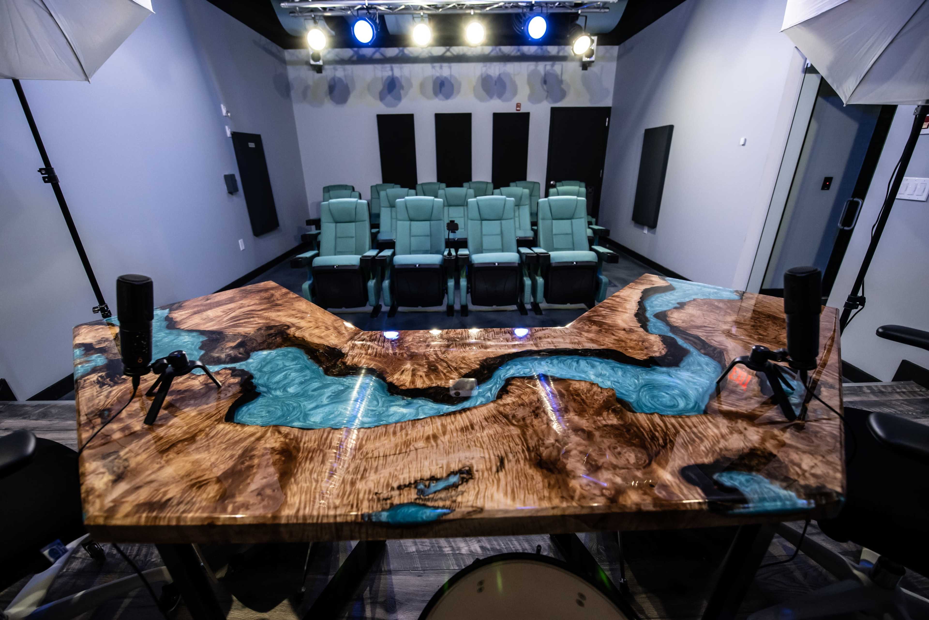 A modern interior featuring a sleek wooden table with turquoise resin inlays and a row of green reclining chairs positioned in front of a stage with spotlights.