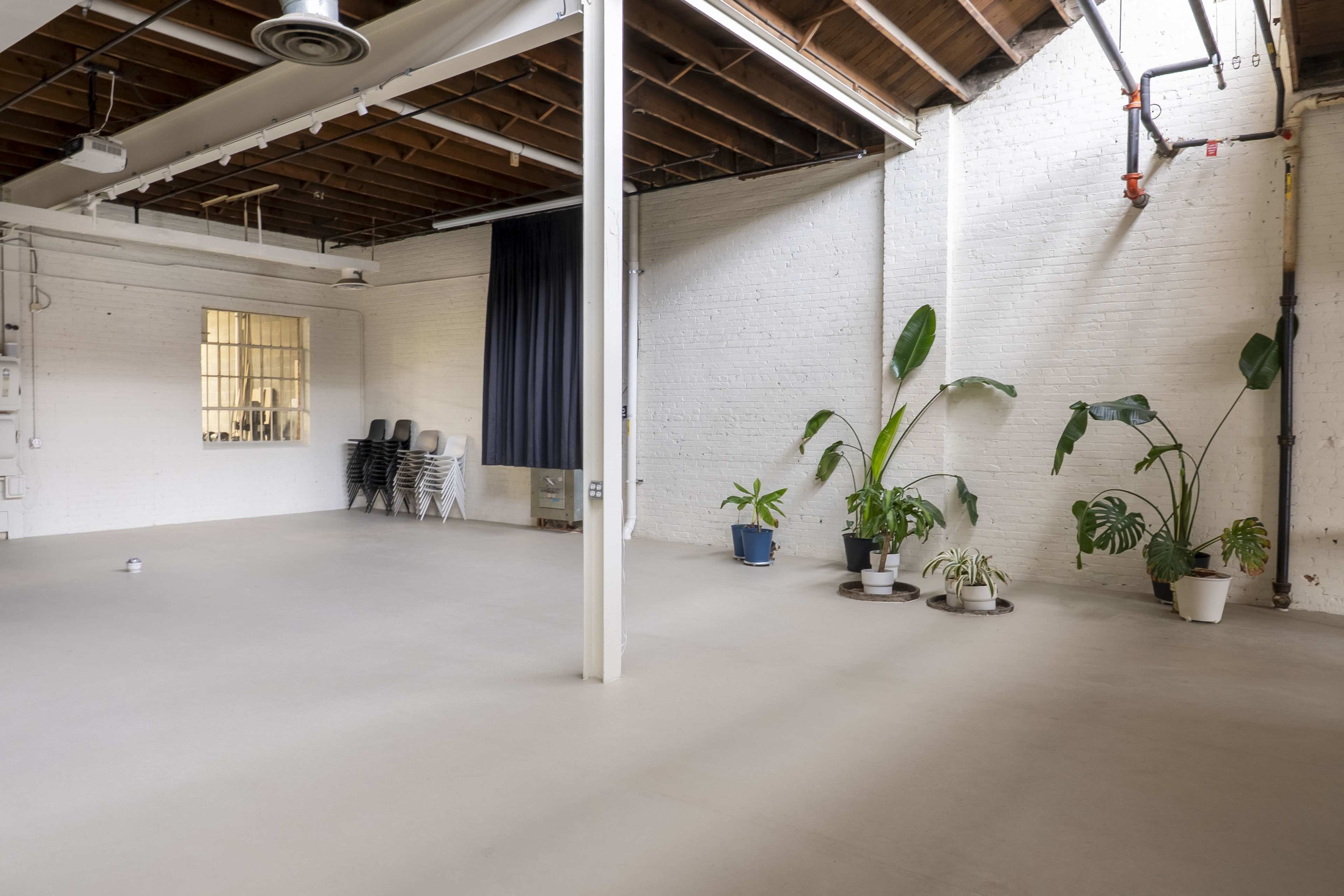 The image shows a spacious, empty room with a polished concrete floor, white brick walls, and a few potted plants along one side.