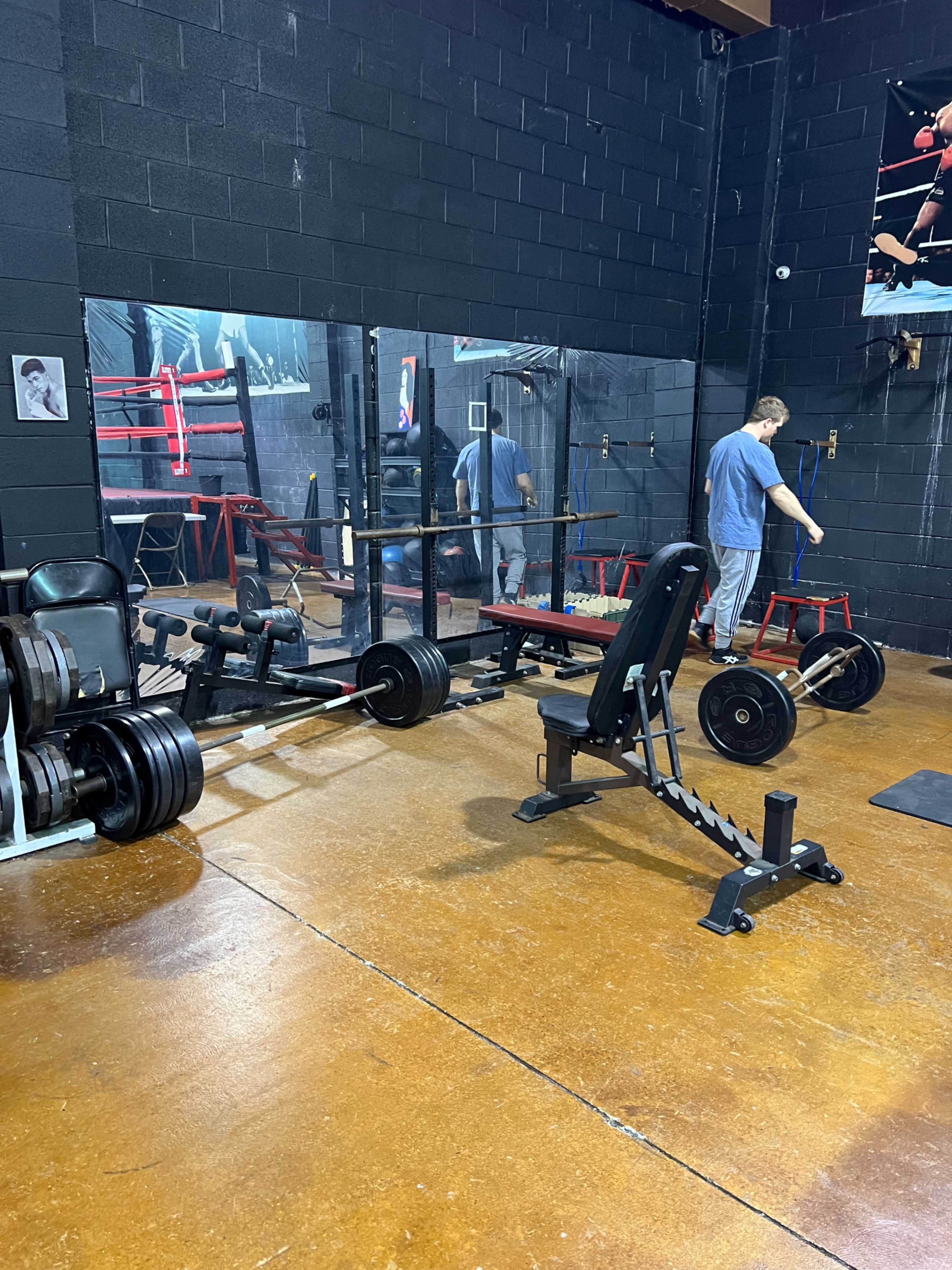 The image shows a gym with various weightlifting equipment, including a barbell and a weight bench, and a person adjusting a piece of equipment in front of a mirror.