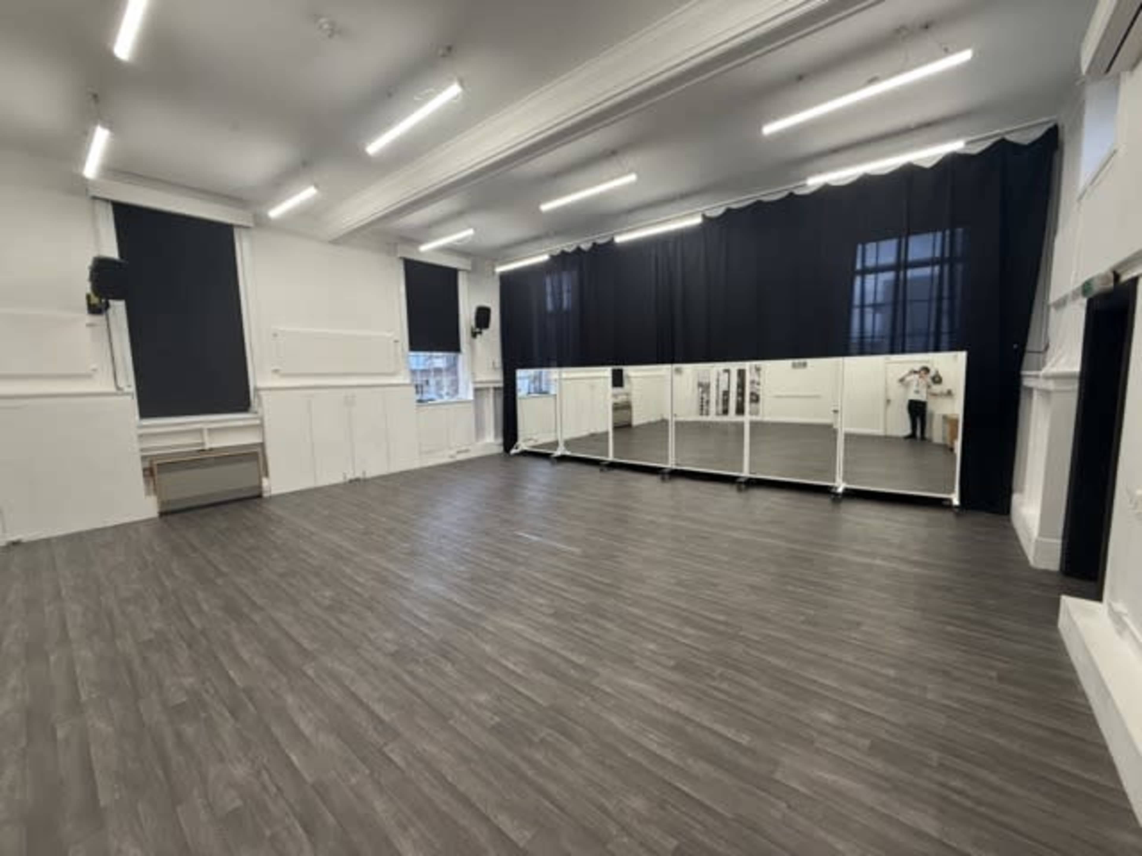 The image shows a spacious dance studio with large mirrors, wooden flooring, and black curtains, illuminated by overhead lights.
