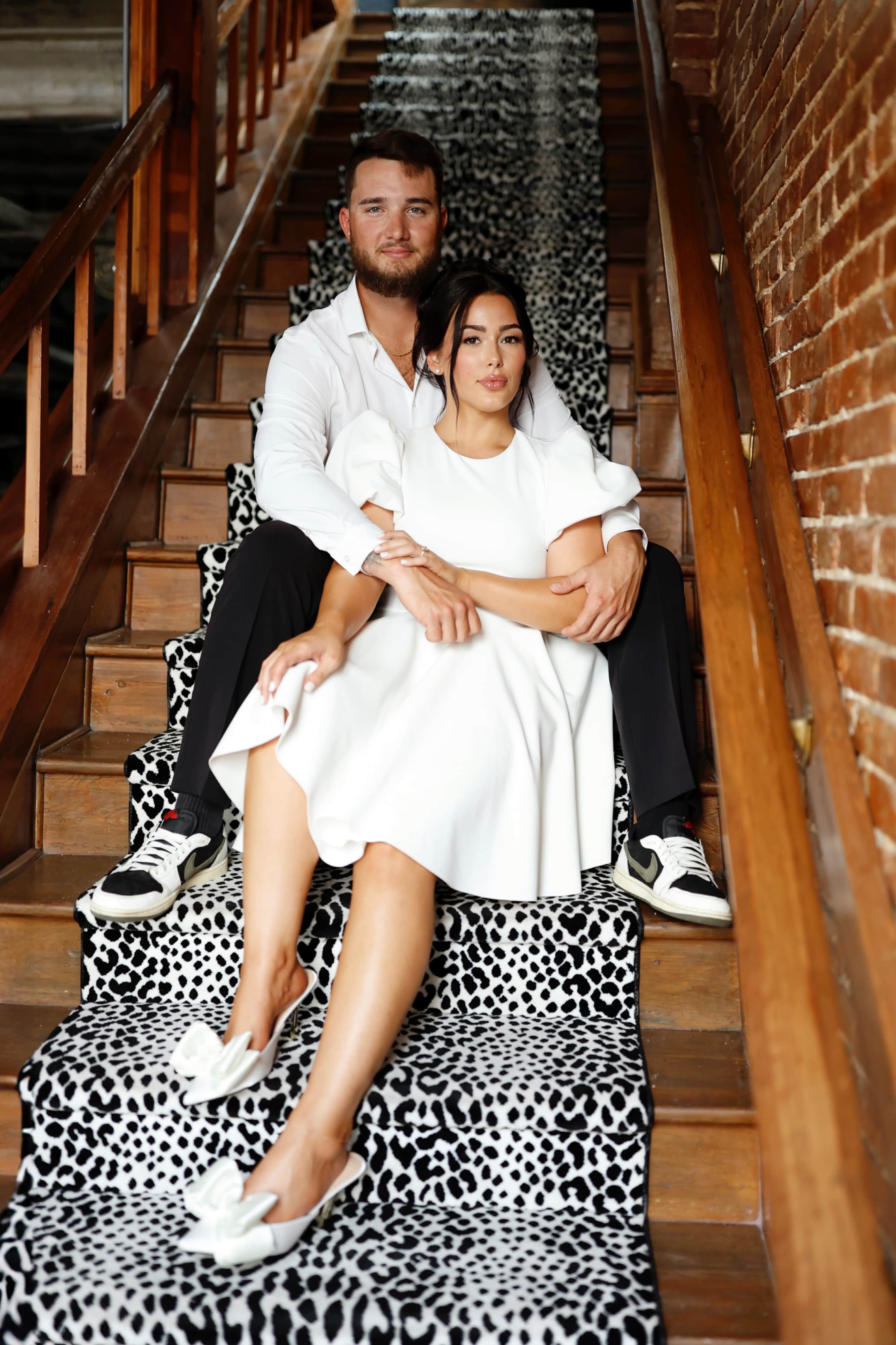 A man in a white shirt and a woman in a white dress sit together on a staircase with a black-and-white patterned runner.
