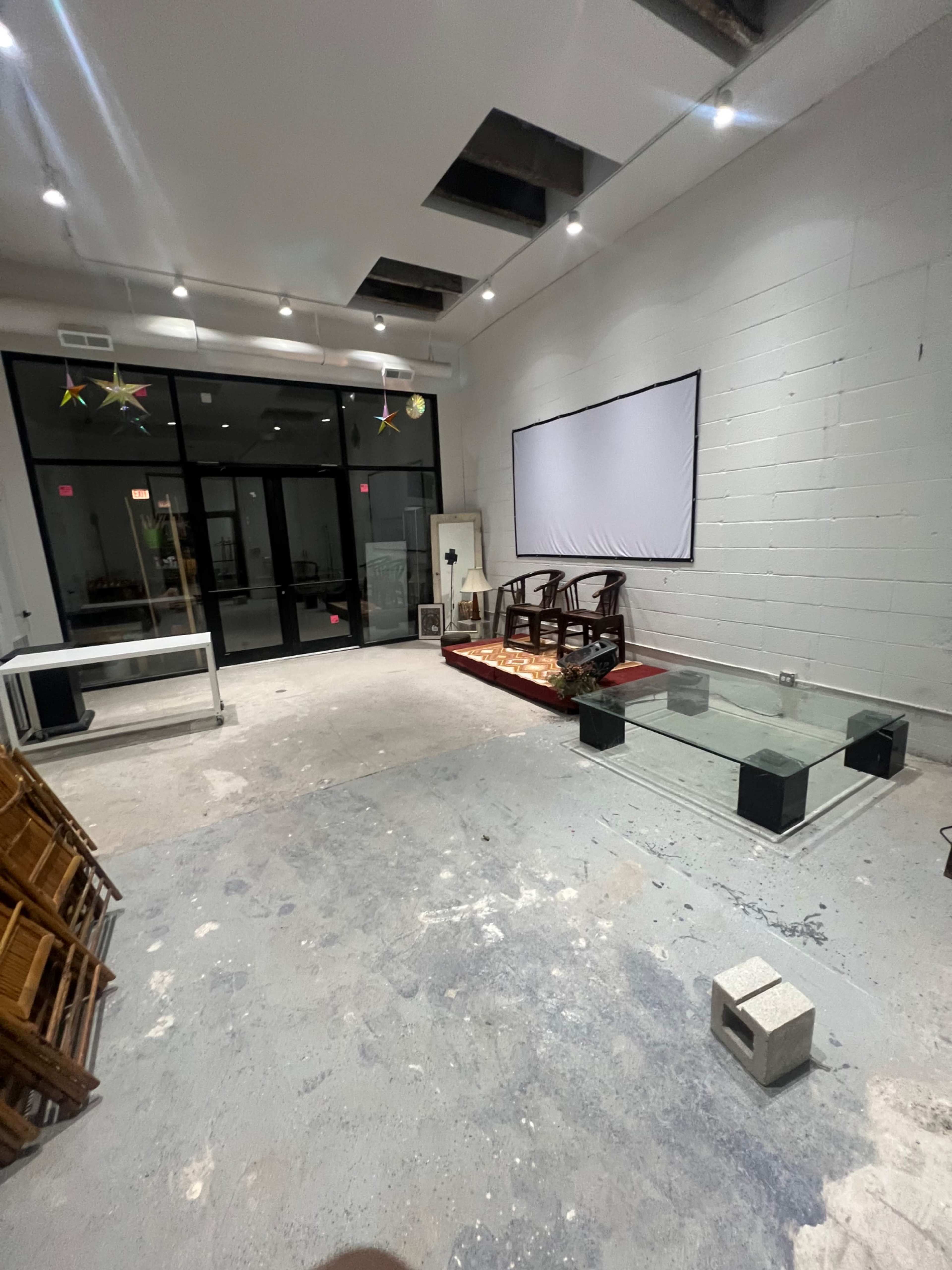 An empty room with concrete floors features some furniture, a white screen mounted on the wall, and large glass windows at the front.