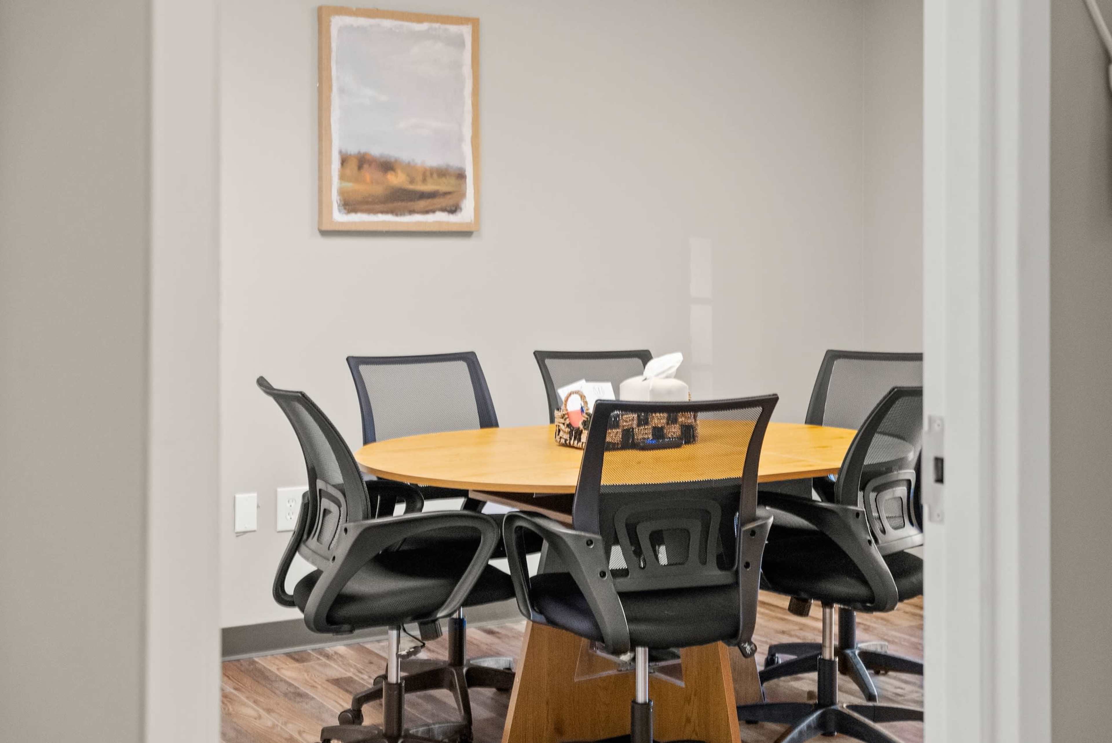 A conference room features a round wooden table surrounded by black ergonomic chairs.