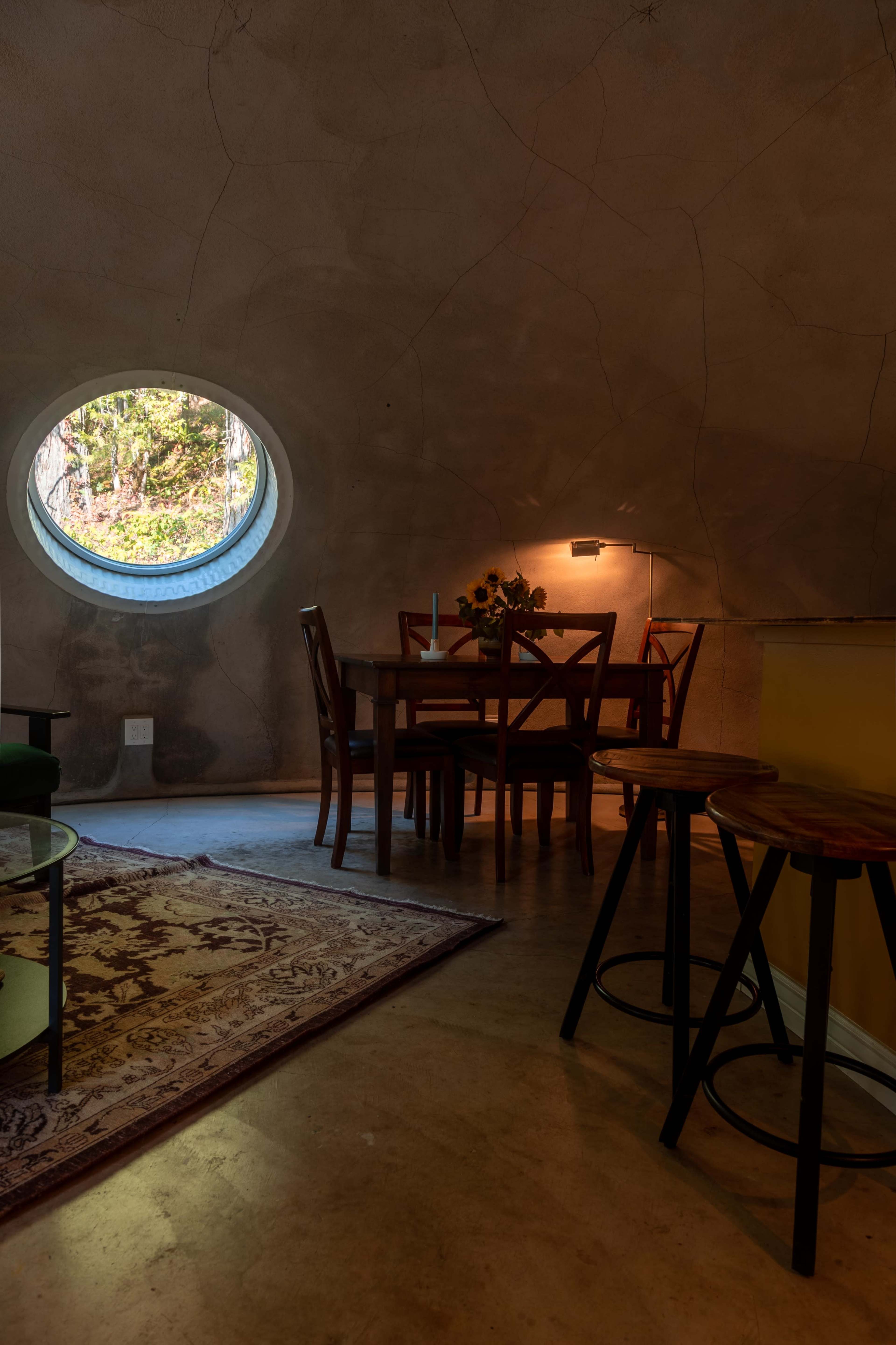 The image shows a round window in an earthen-colored room with a dining table and chairs, illuminated by soft light and featuring a small rug on the floor.