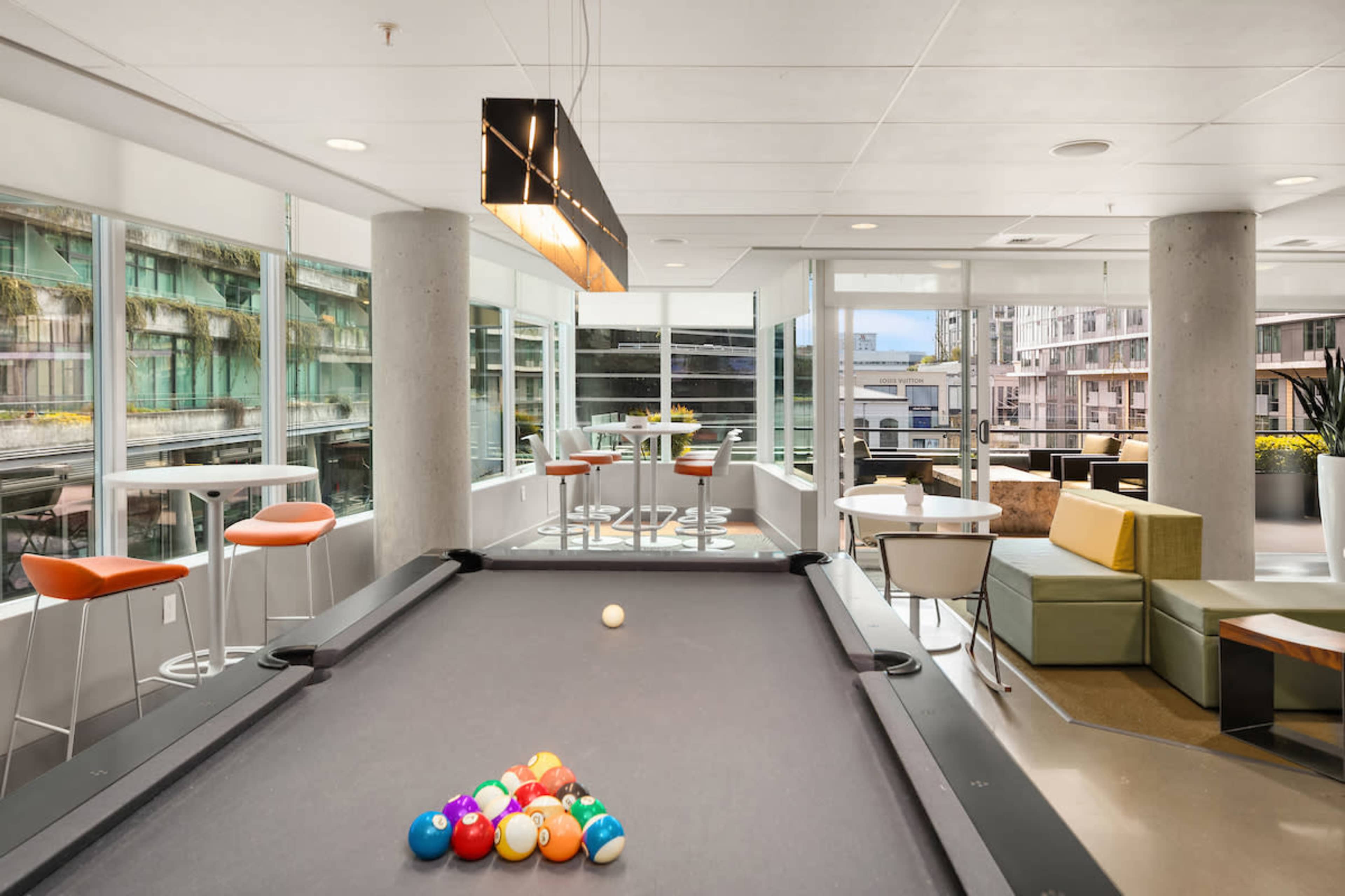 The image shows a billiard table with colorful balls arranged for a game in a modern lounge area filled with large windows and contemporary furniture.