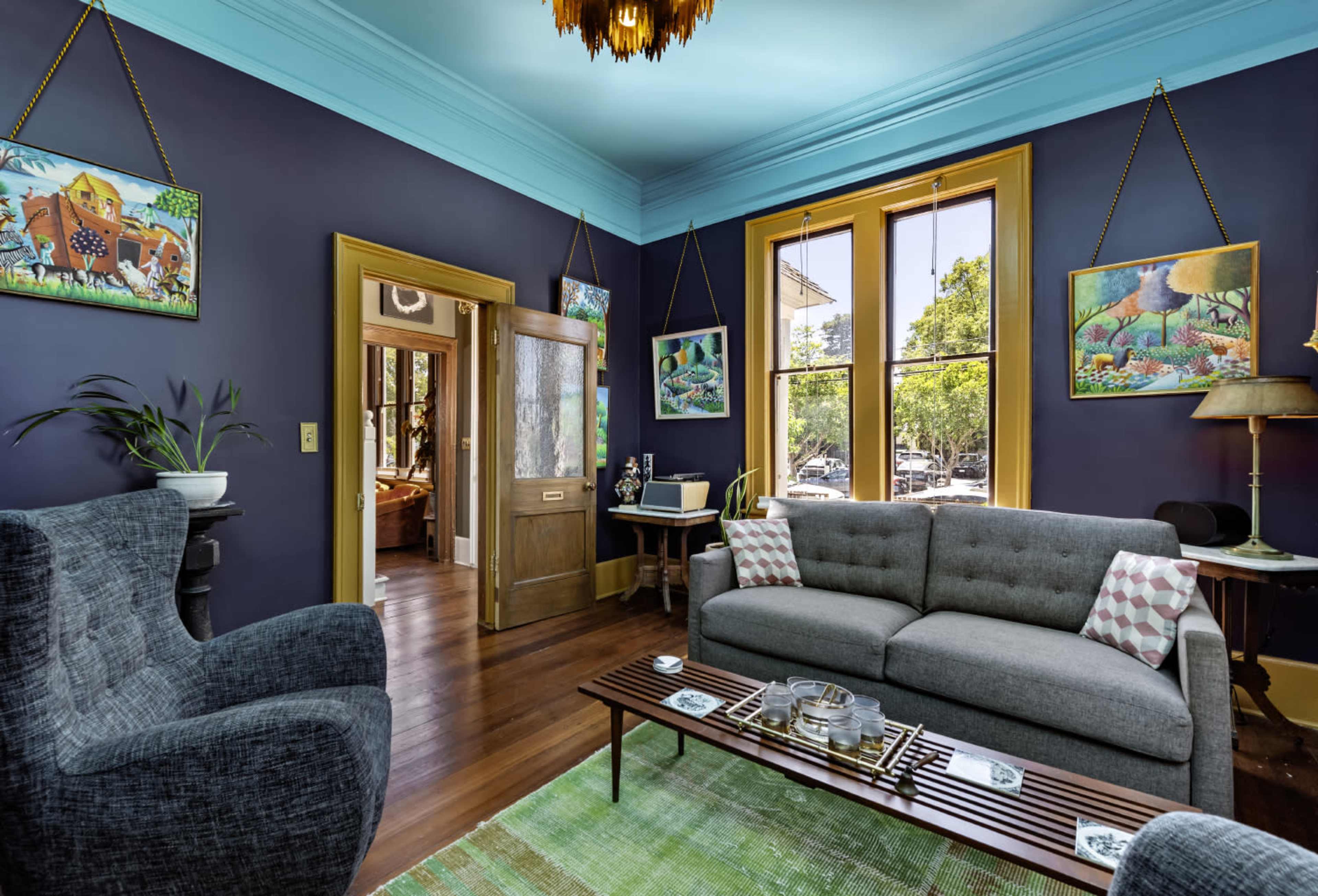 The image shows a colorful living room with dark blue walls, a gray sofa, and large windows that provide natural light.