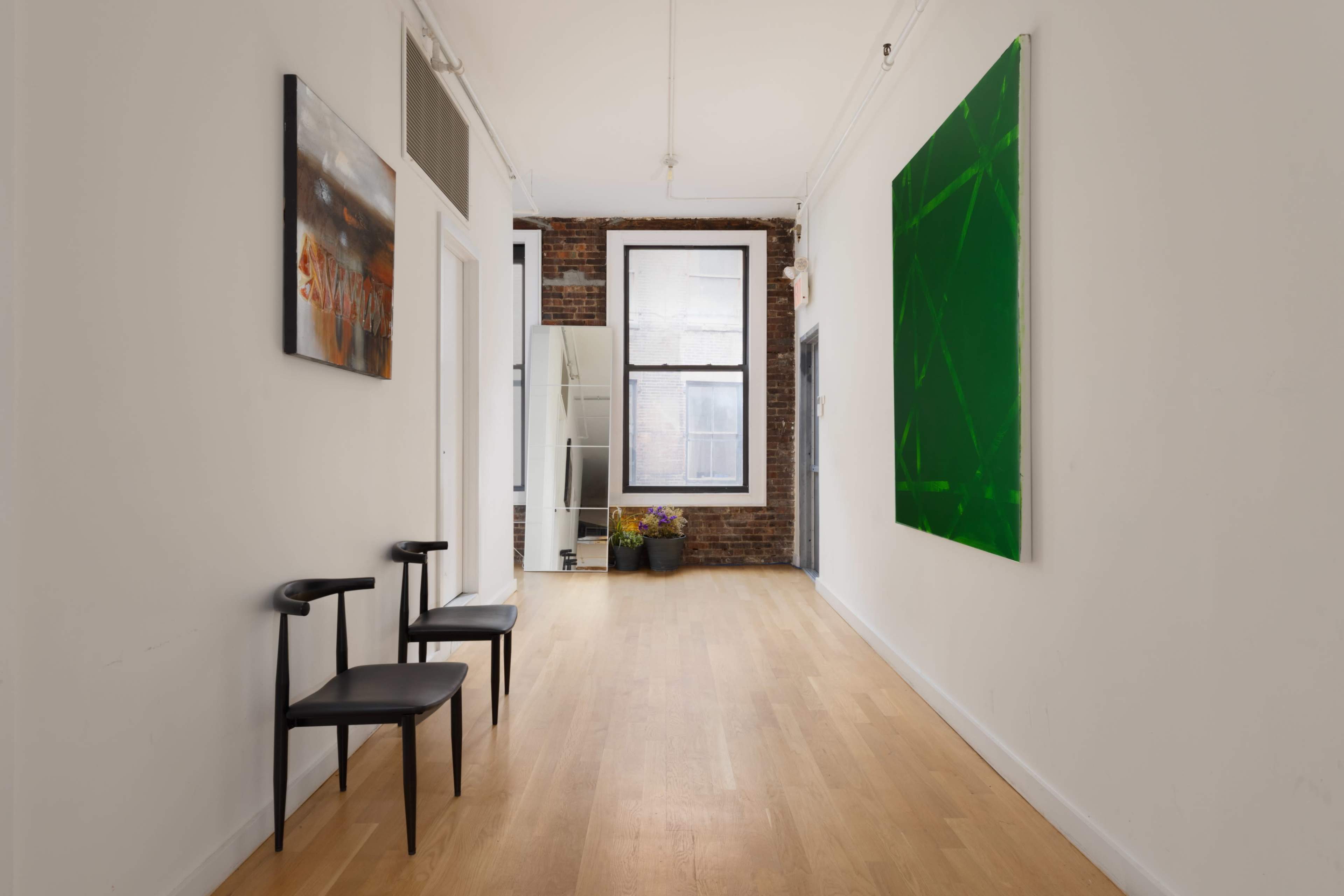 A long, narrow hallway featuring two black chairs, a large green painting on the wall, and exposed brick along one side.