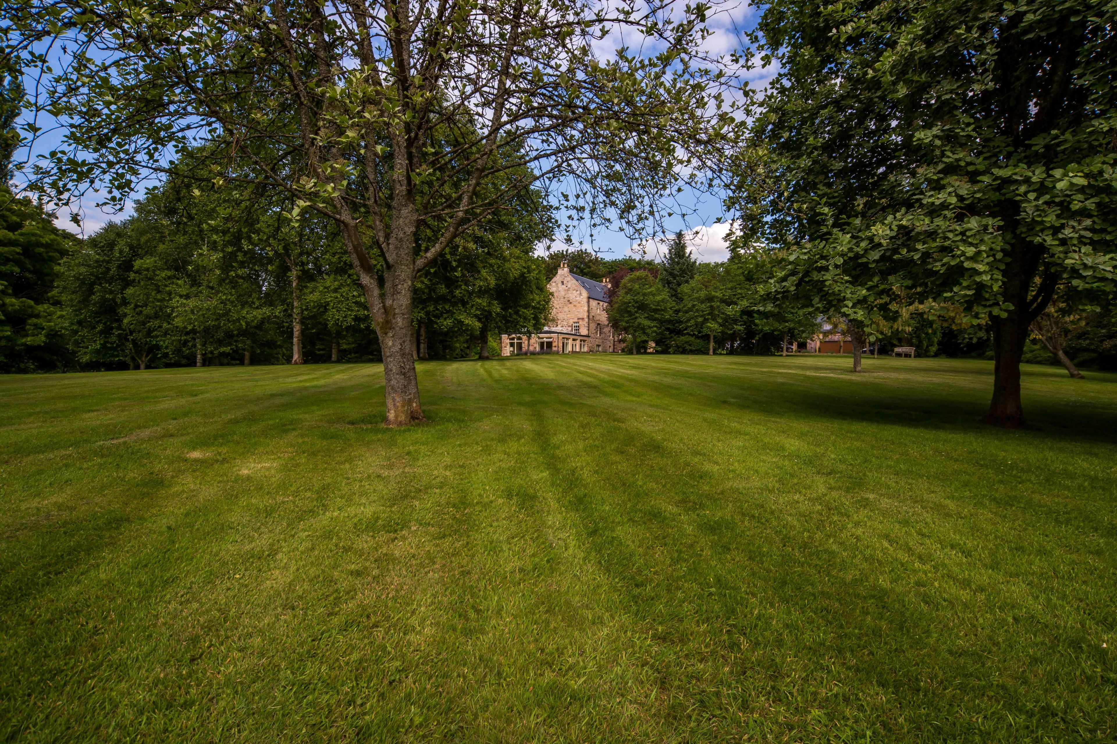 A large, open lawn with manicured grass leads to a stone house partially obscured by trees.
