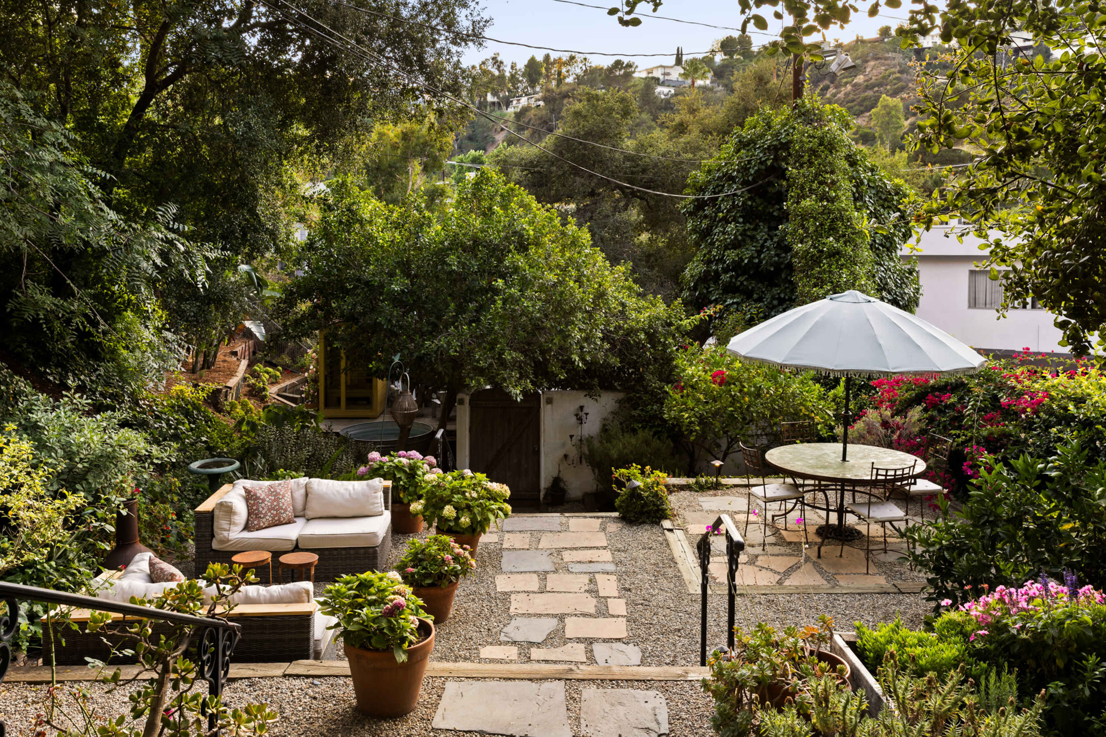 The image shows a landscaped garden with stone pathways, potted plants, a seating area with a sofa, a round dining table under an umbrella, and surrounding greenery.