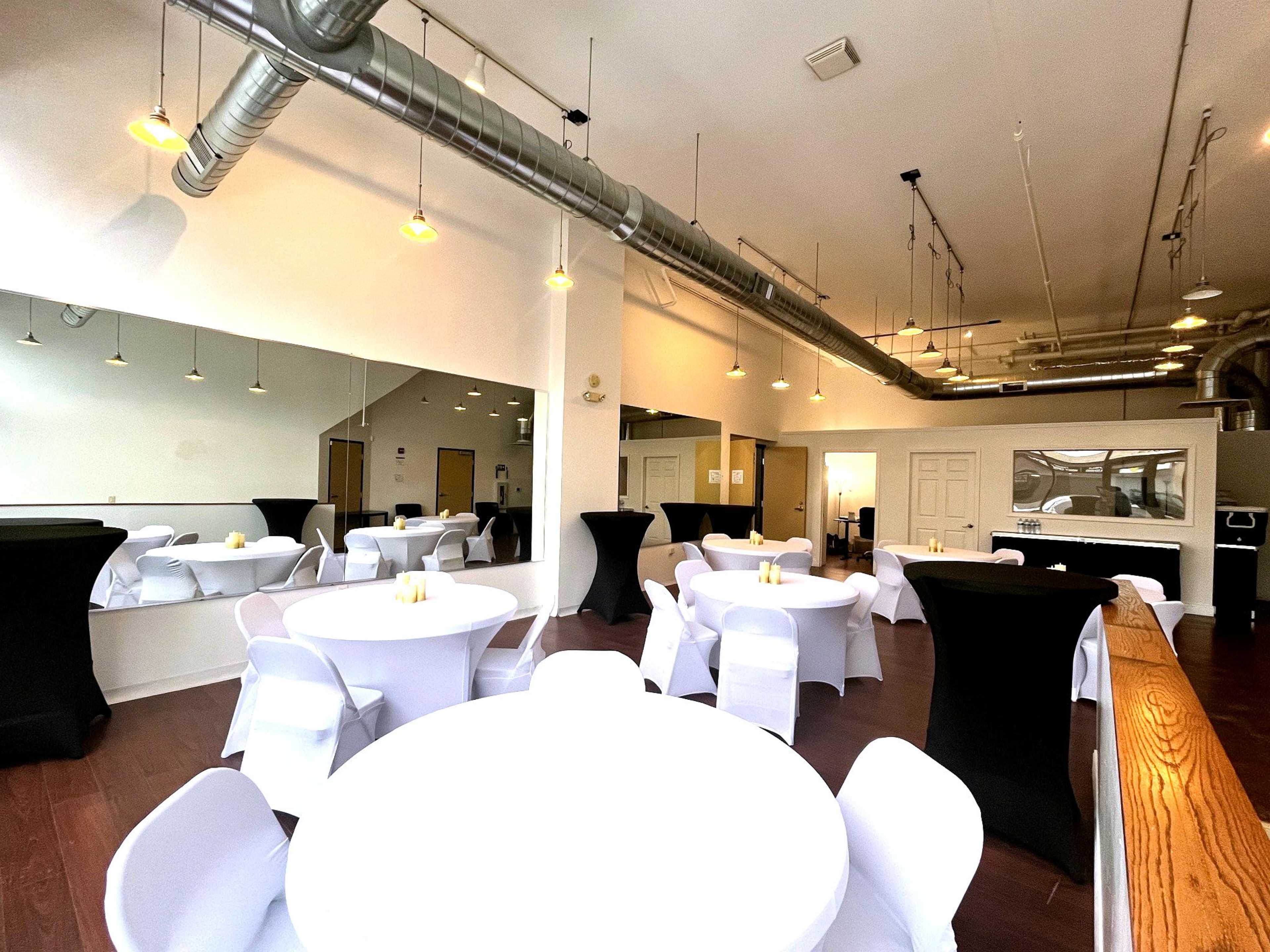The image shows a spacious event venue with several white and black tables set up, featuring a large mirror and industrial-style ceiling fixtures.