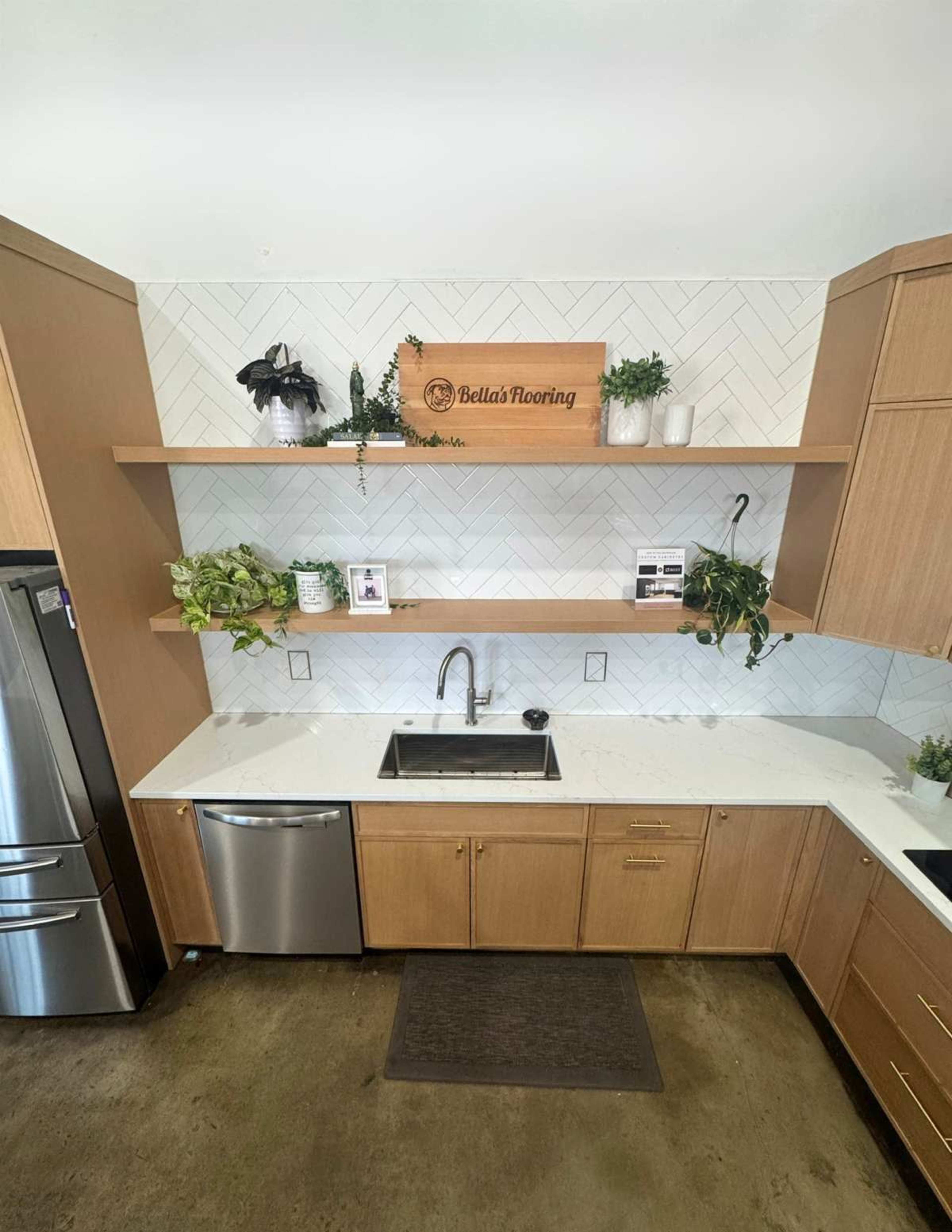 The image shows a modern kitchen with wooden cabinets, a stainless steel refrigerator, a stainless steel sink, and open shelves displaying plants and decorative items.