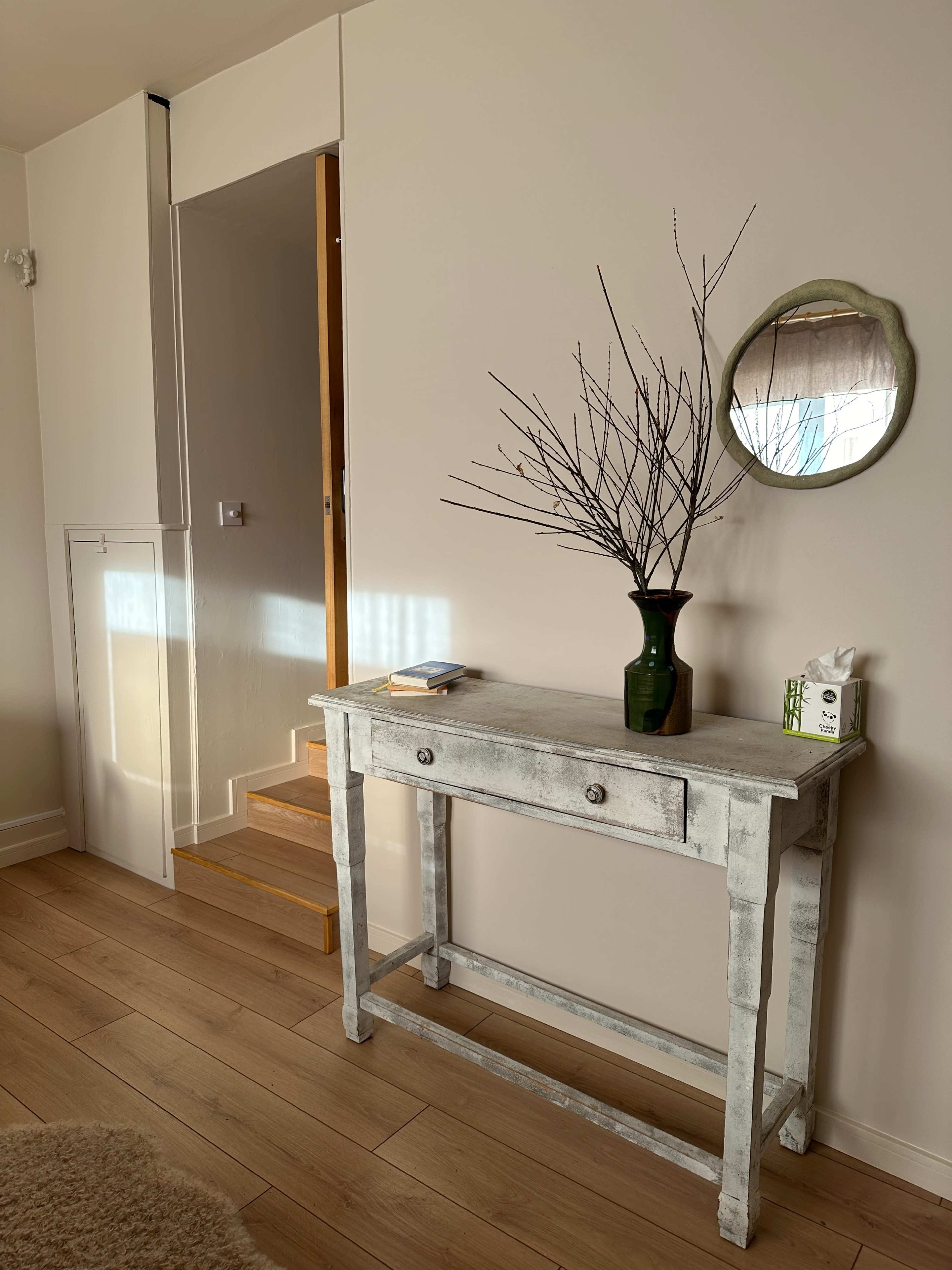 A wooden console table with a single drawer is positioned against a wall, adorned with a green vase and a branch arrangement, near a staircase leading upwards.