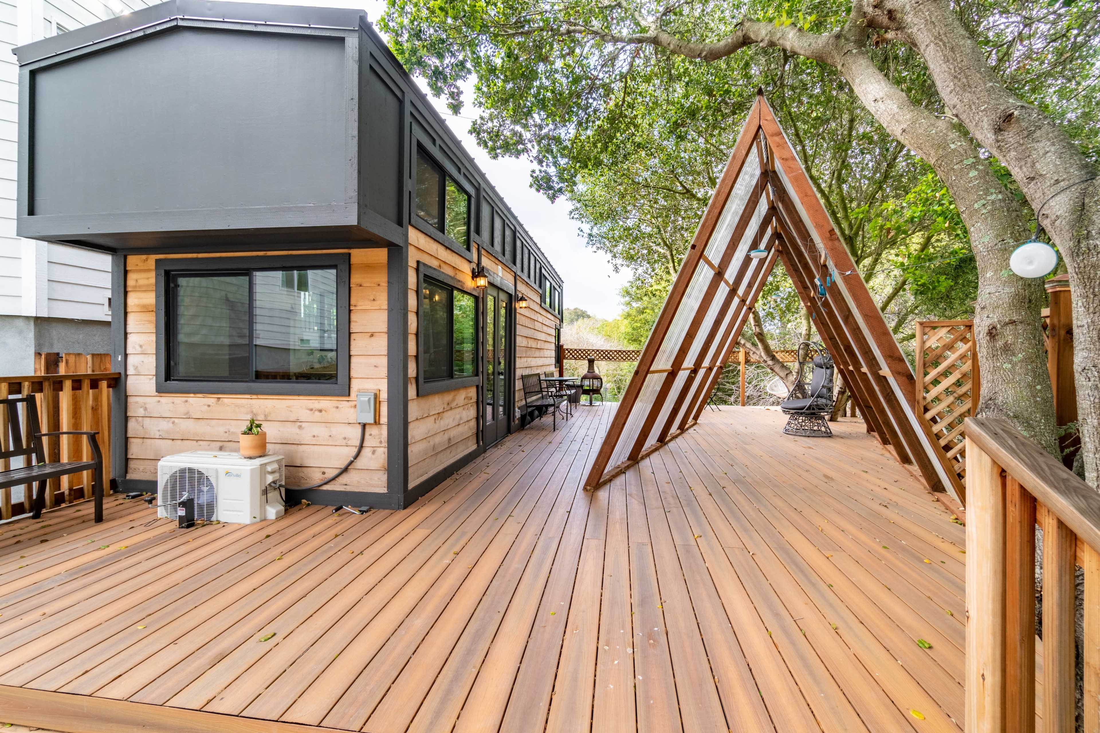 The image shows a modern wooden deck with a cabin-style structure and a triangular wooden feature beside it, surrounded by trees.
