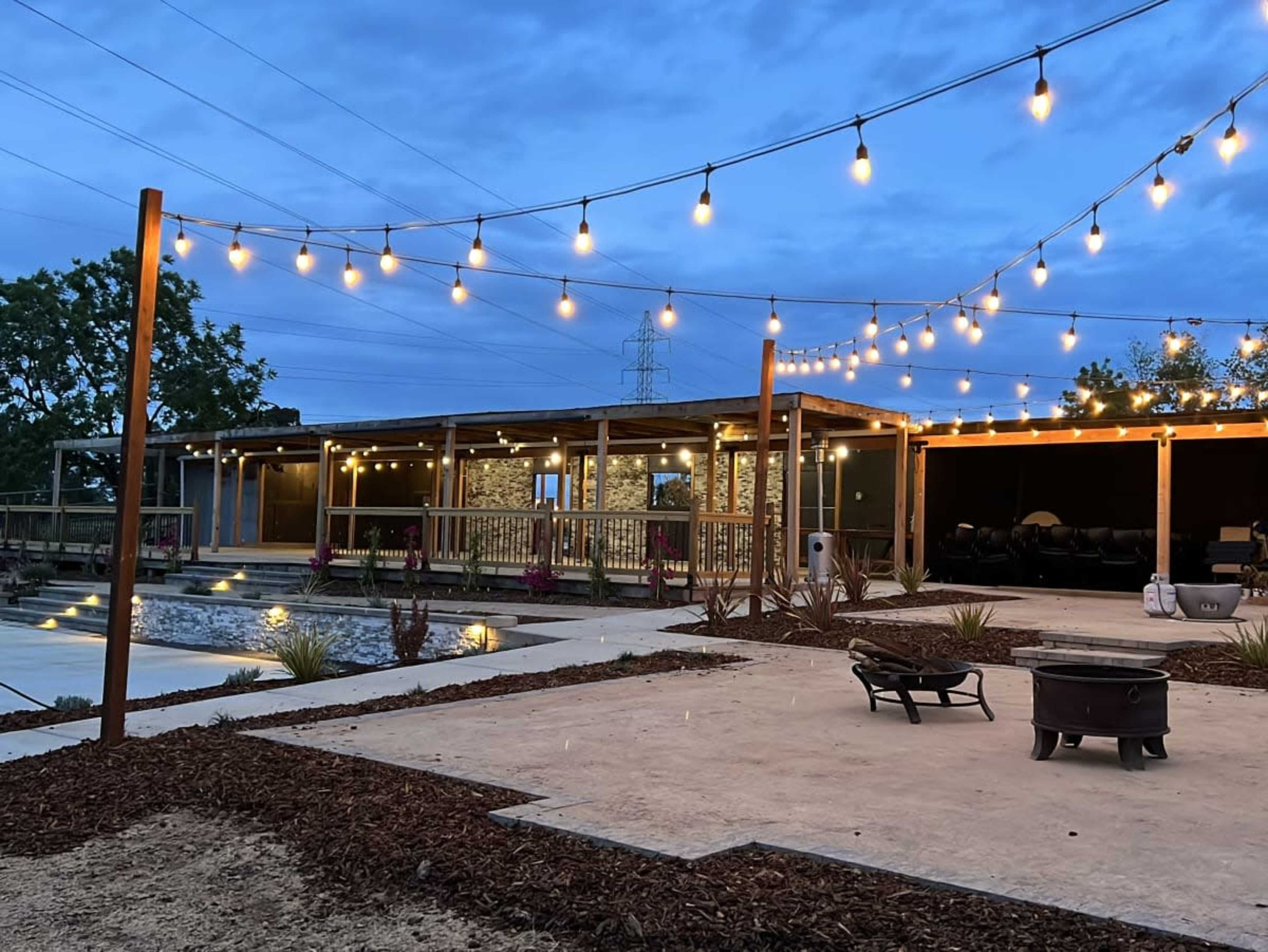 The scene shows a patio area with hanging string lights, a pool, and a modern building surrounded by landscaped gravel and plants under a twilight sky.