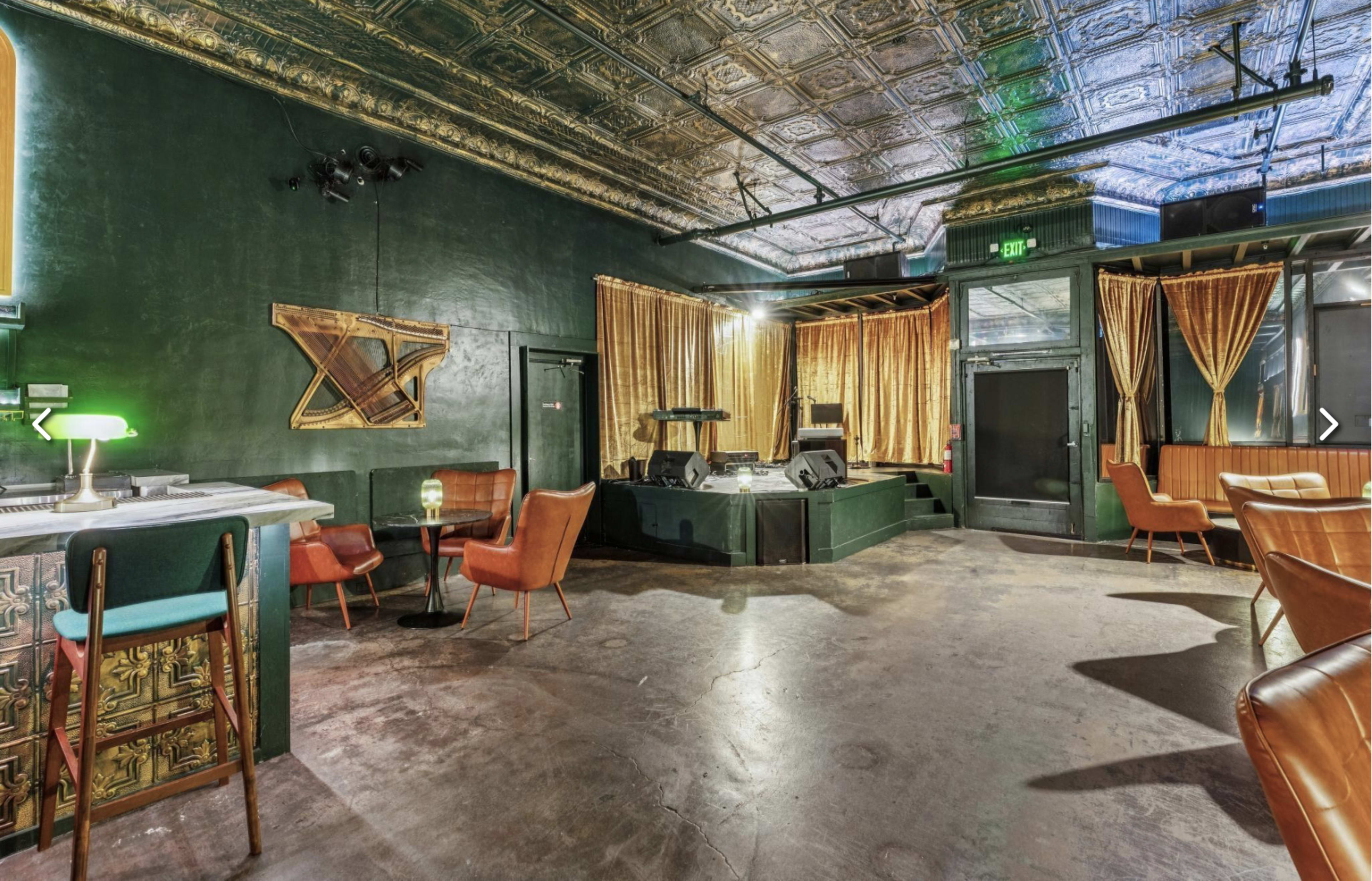 The image shows a dimly lit venue with a raised stage, velvet curtains, and a bar area featuring several chairs and tables.